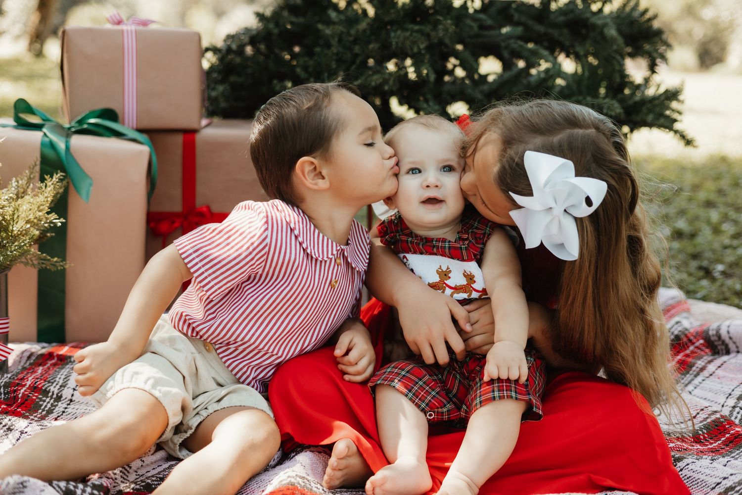 Two older siblings kissing a baby on the cheeks during a festive Christmas photo session with gifts and a tree.