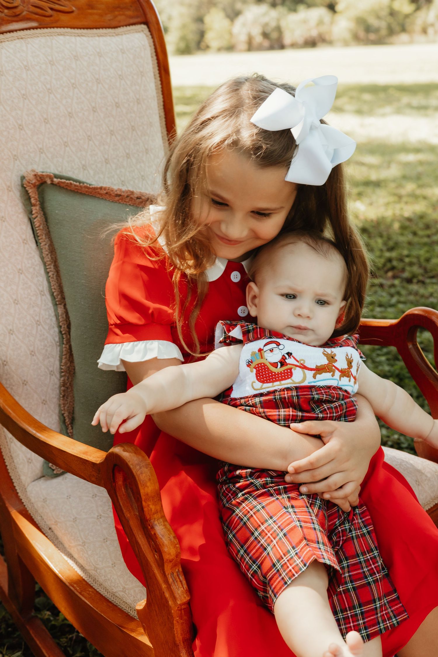Girl in red dress with white bow holds baby in Christmas plaid outfit in a rocking chair outdoors.