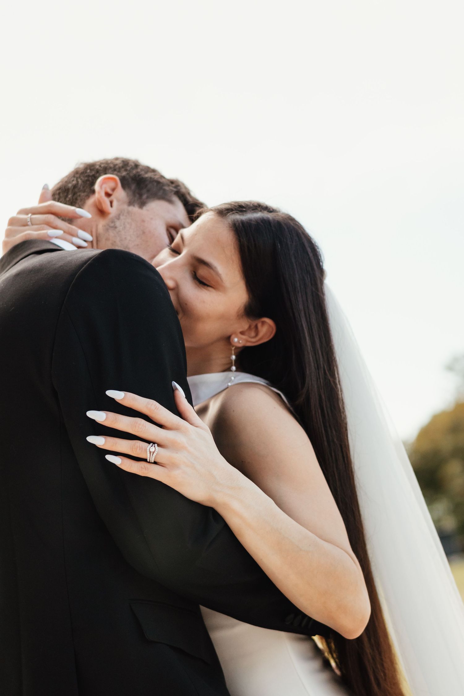 Bride and groom share a romantic kiss, bride wearing a veil and diamond ring, embracing in an outdoor wedding setting.