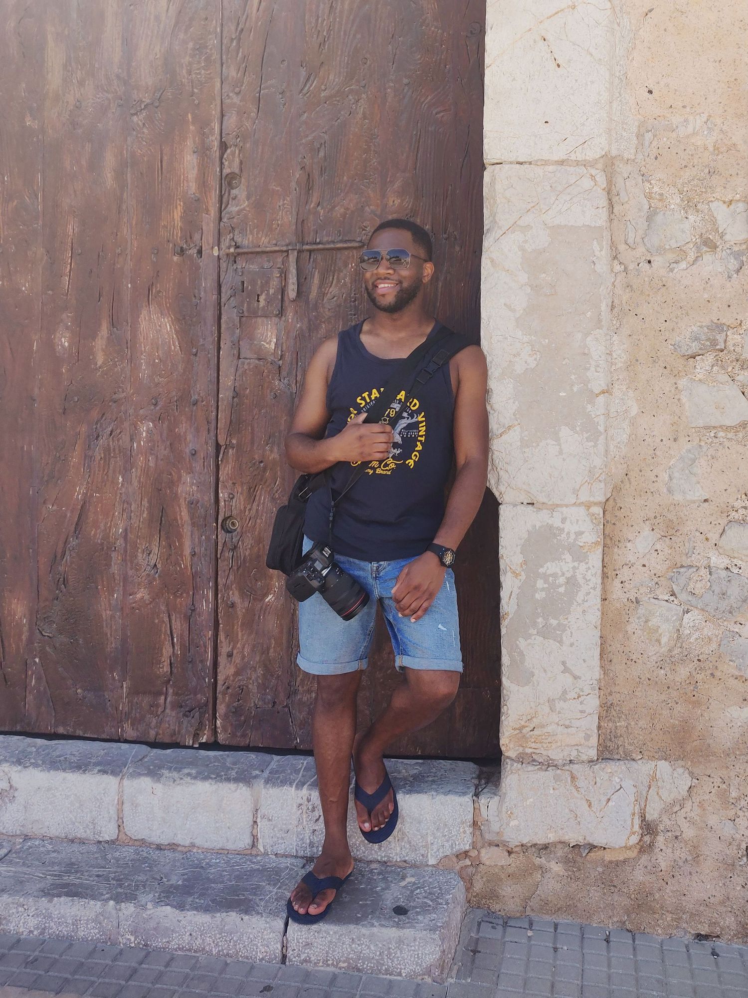 Person in casual summer attire leaning against rustic wooden door of historic building.