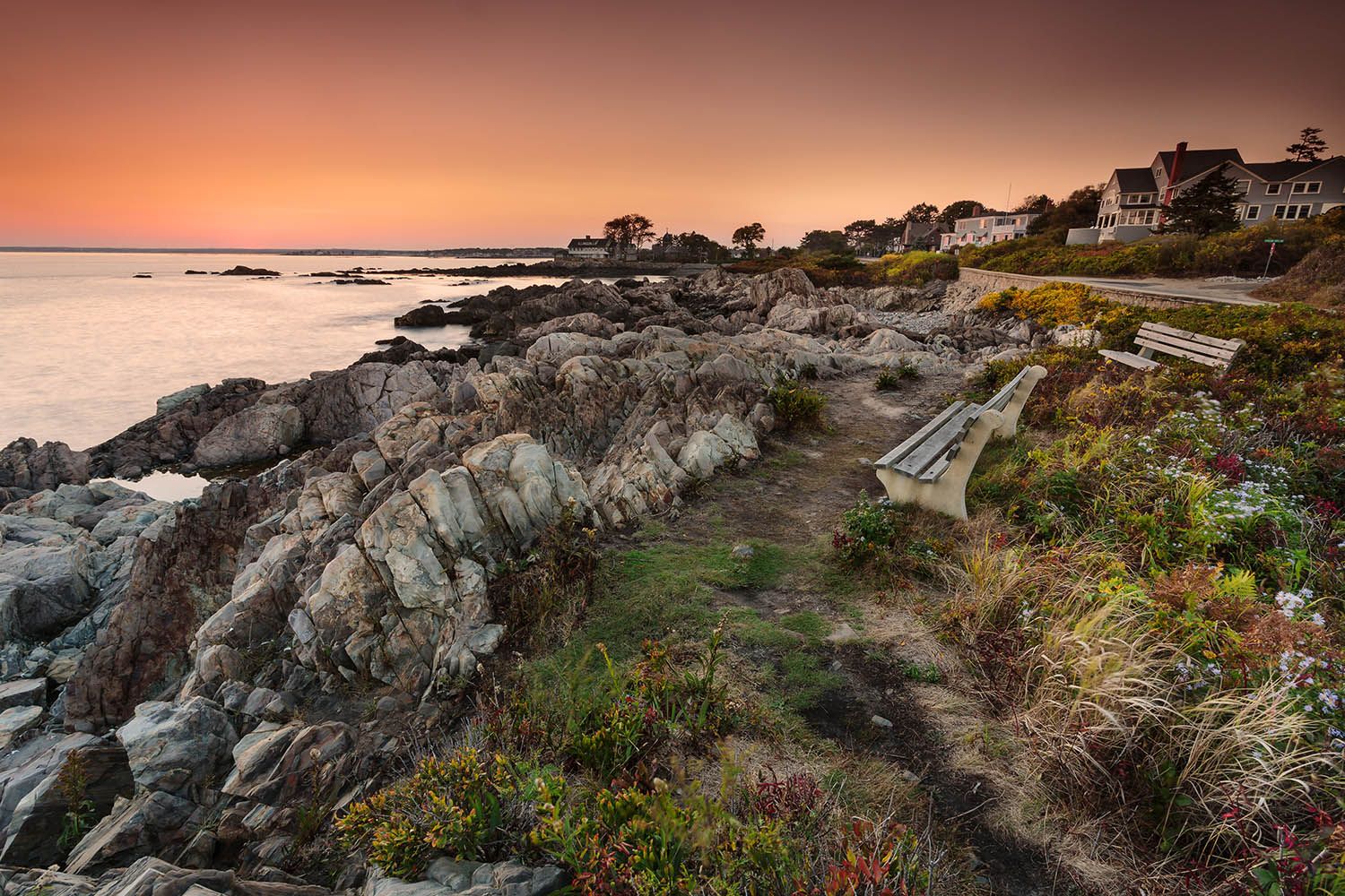 Color landscape photograph showing the coast of Maine near Kennebunkport at sunset with beach in the background and bench in the foreground.