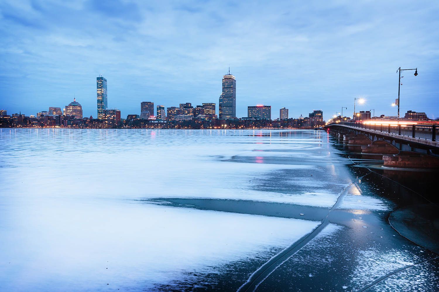 Winter landscape photograph in color of the Charles Rive frozen and a dark and moody sky over the Boston city skyline with bridge on right