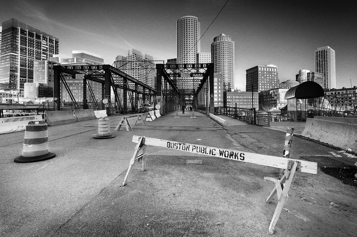 Black and white cityscape photography of Boston taken from the Seaport District and police barrier in the foreground with derelict bridge