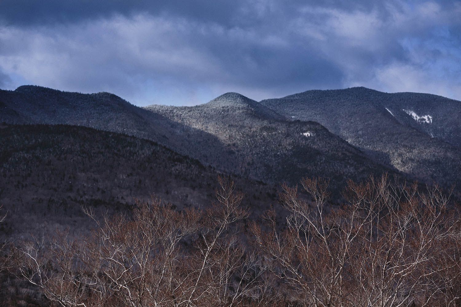 Color landscape photograph of the White Mountains NH during the winter with snow starting to build on the peaks and bare tress in foreground