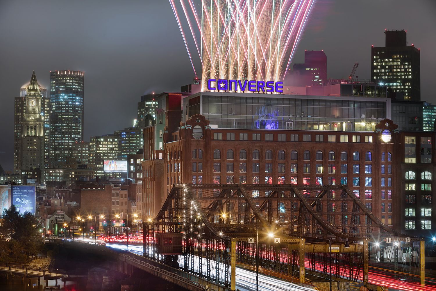 BOSTON ARCHITECTURAL PHOTOGRAPHERS CAPTURE CONVERSE HQ AT NIGHT WITH FIREWORKS GOING OFF AND BOSTON CITY IN THE BACKGROUND