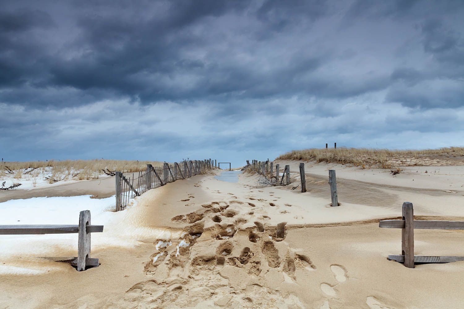 Winter beach landscape photograph in color of pathway leading up the dunes and to the beach. Snow is settling on the sand the sky is dark