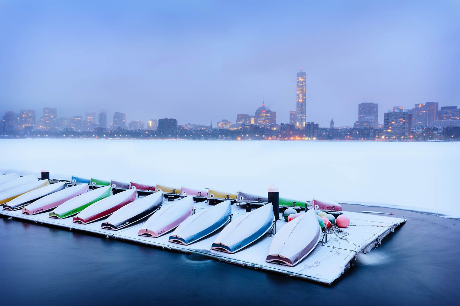 Color landscape photograph of the Charles River during the winter with Boston skyline in the back boats on the pontoon and covered in snow
