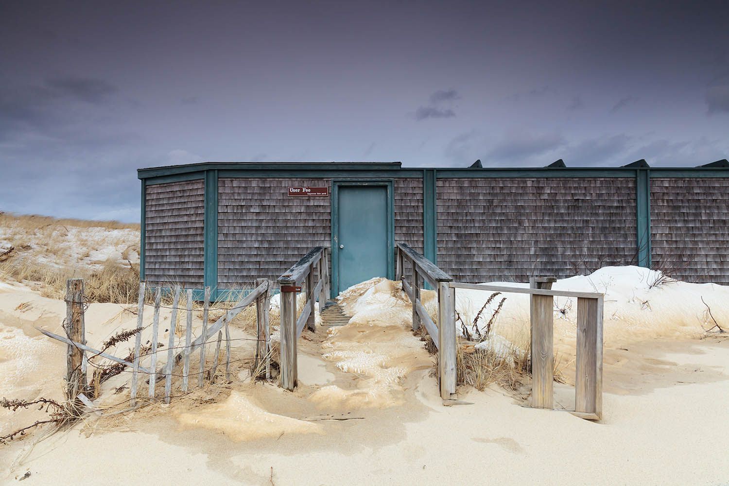 Winter beach landscape photograph in color of small facilities hut that has been closed up for the year the sand and snow have blown over