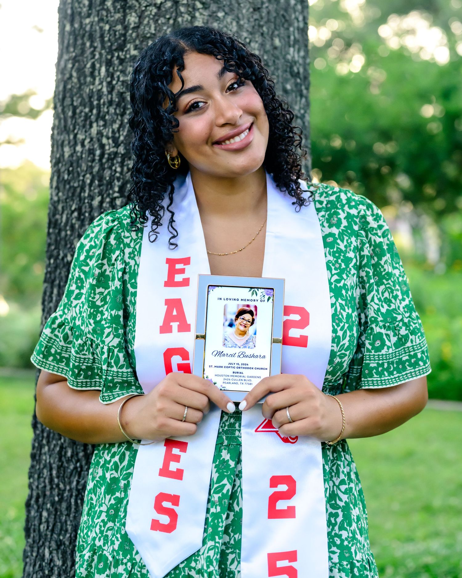 Graduate in green floral dress with white graduation stole and ID card stands by tree in outdoor setting.