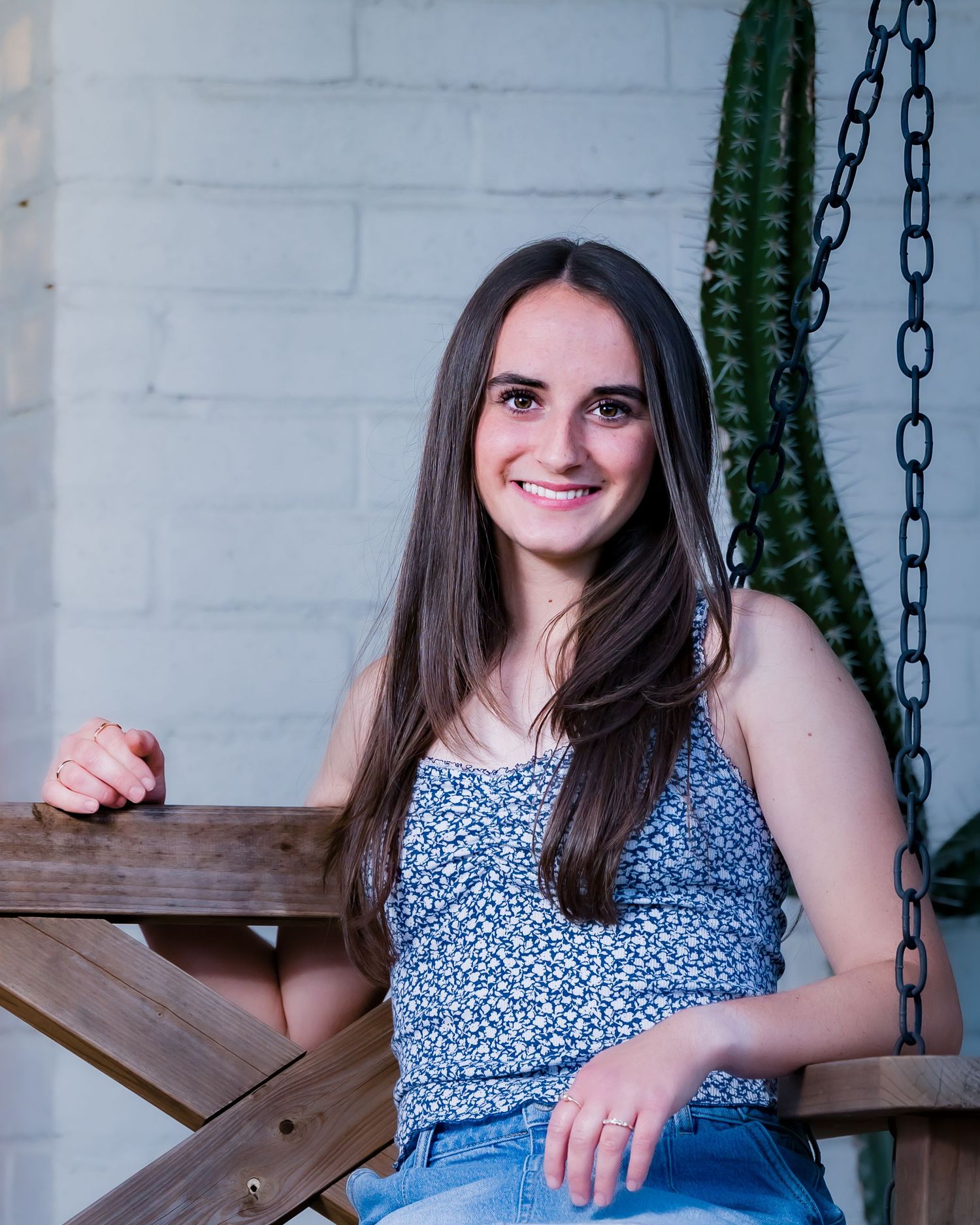 Young woman with long brown hair smiling while sitting on a wooden swing bench outdoors.
