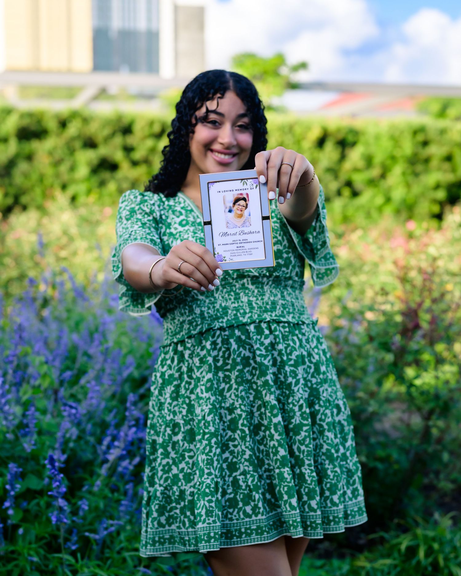 Someone in a green floral dress stands in a garden with purple flowers displaying a card on a sunny day.
