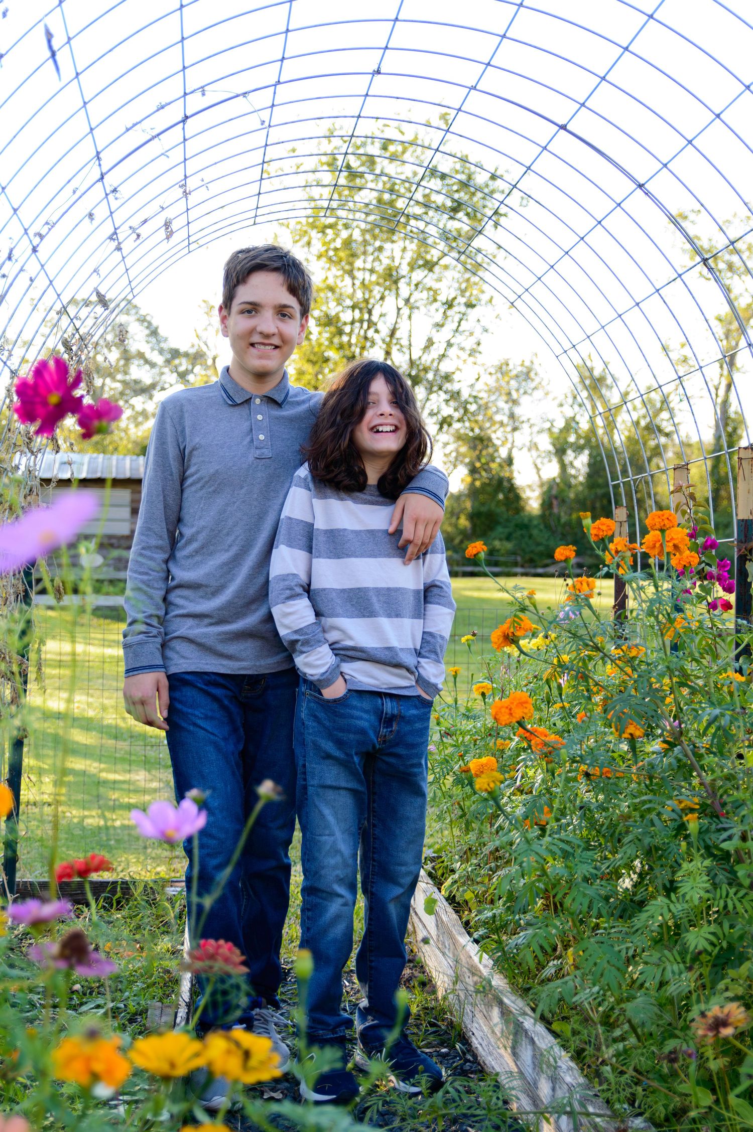 Two young people stand together in a greenhouse walkway surrounded by colorful flowers and garden plants.