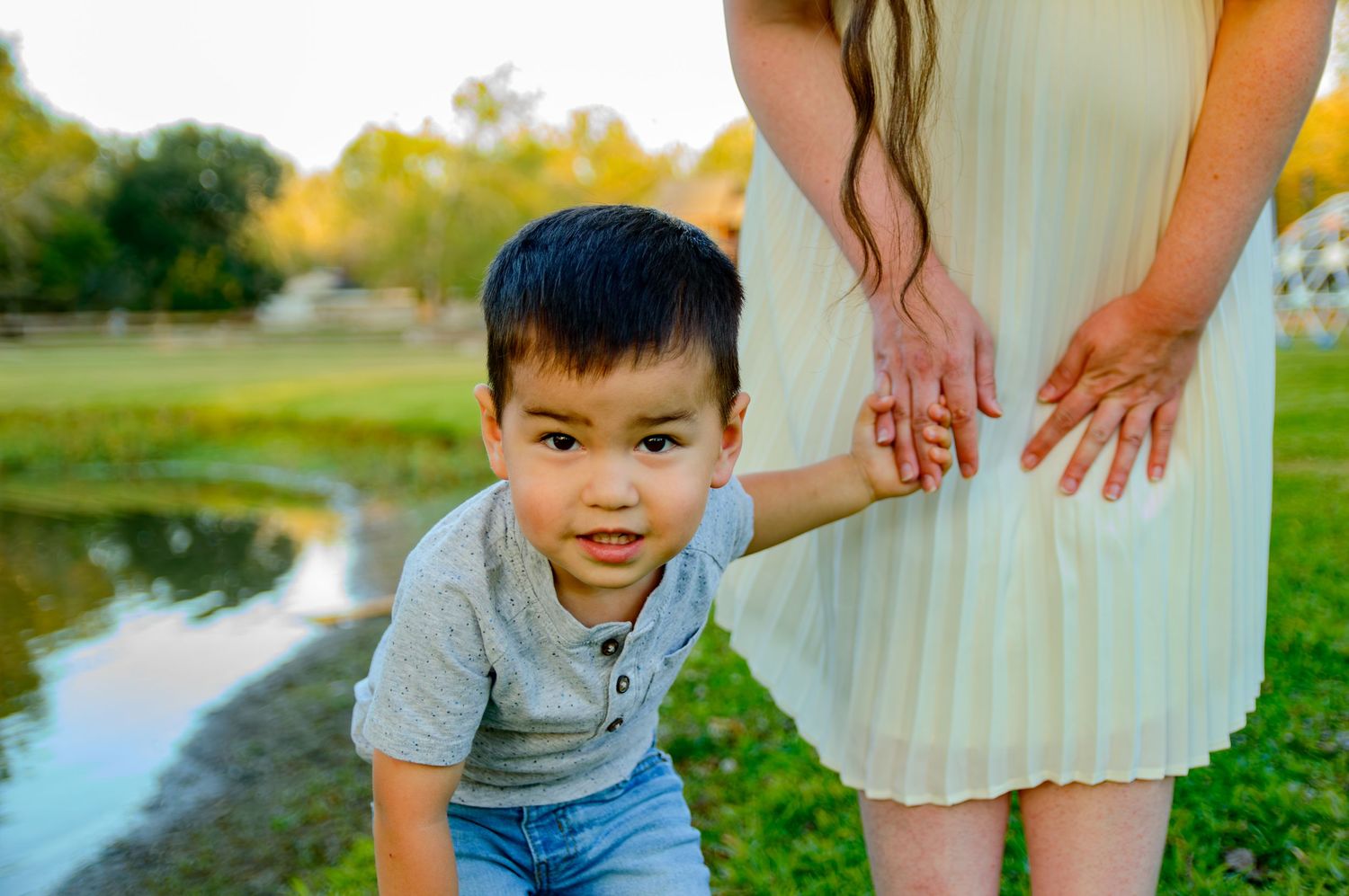 Child in grey shirt smiles while playing outdoors near a pond on a sunny autumn day.