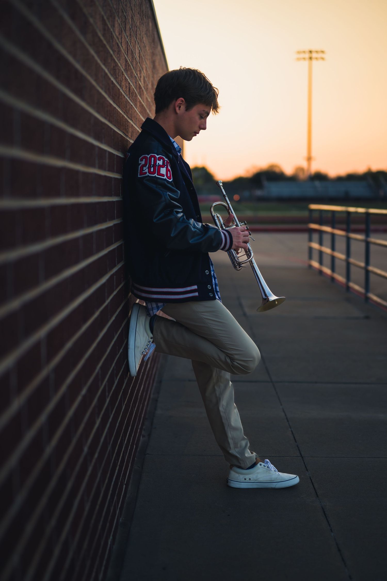 Person sits against brick wall at sunset wearing varsity jacket and holding a smartphone in moody lighting.