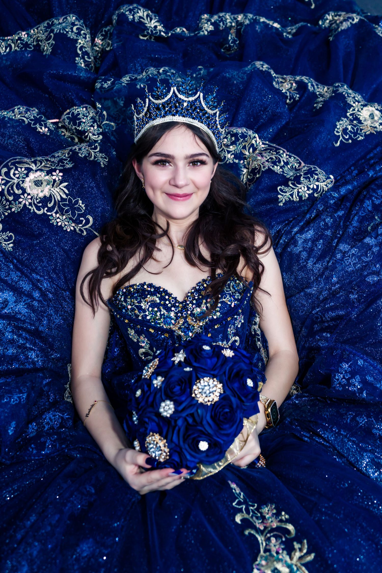 A portrait against a deep blue velvet backdrop wearing a sparkly navy blue gown and ornate crown.