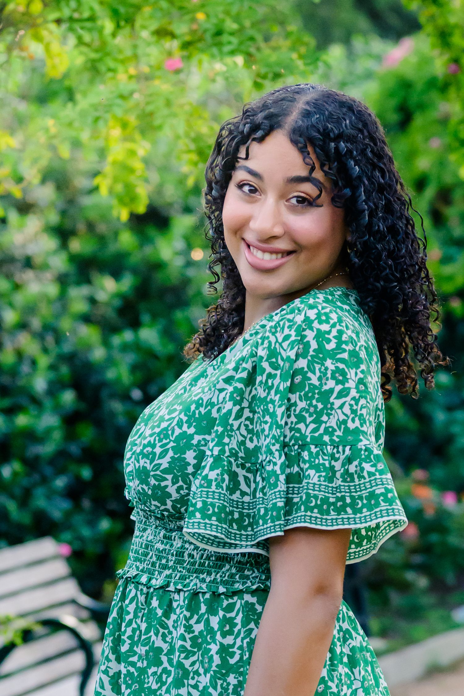 A person in a green and white floral dress with curly hair poses outdoors against a garden backdrop.