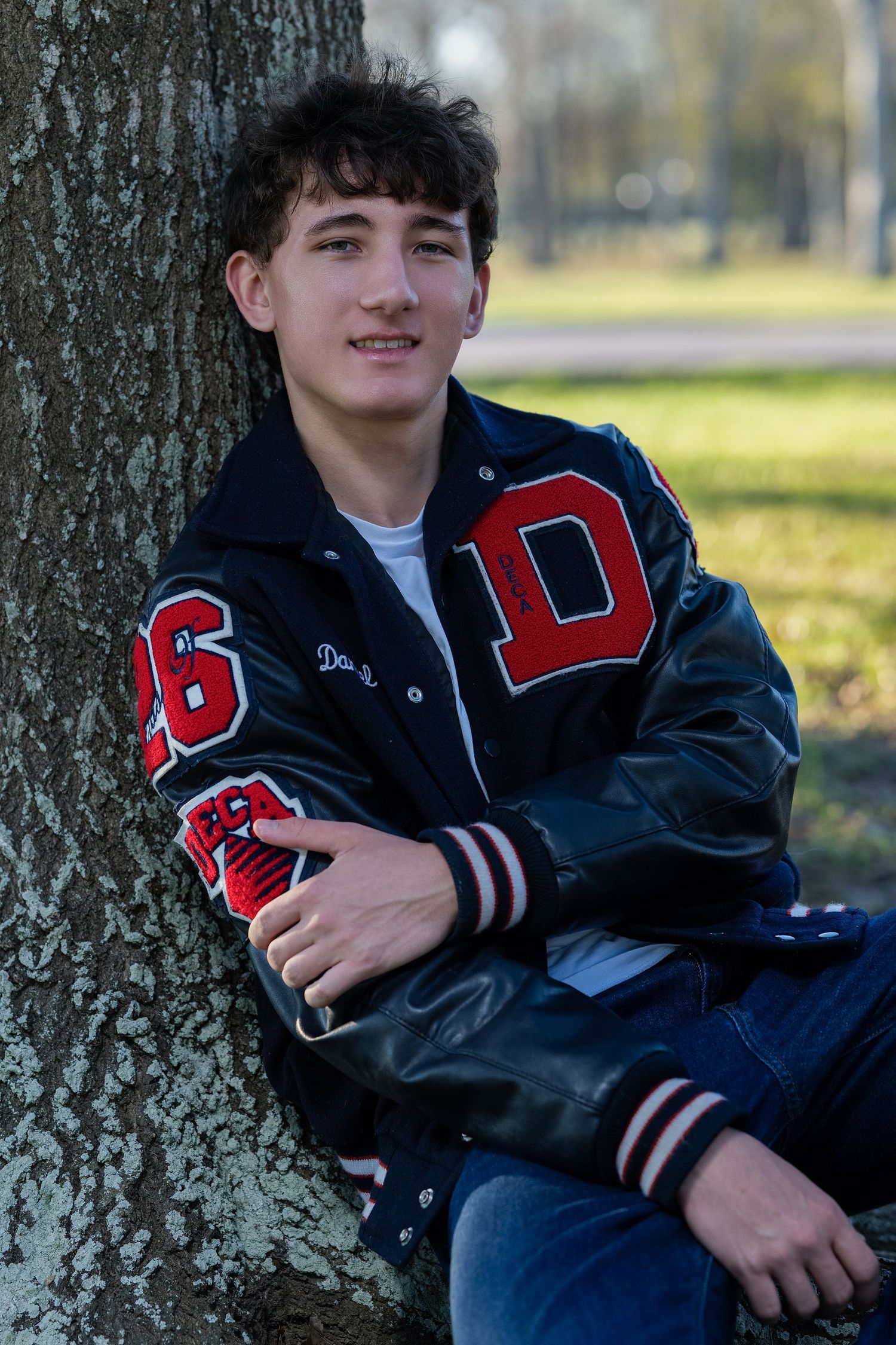 A student in a varsity letterman jacket leans against a tree trunk in an outdoor autumn setting.