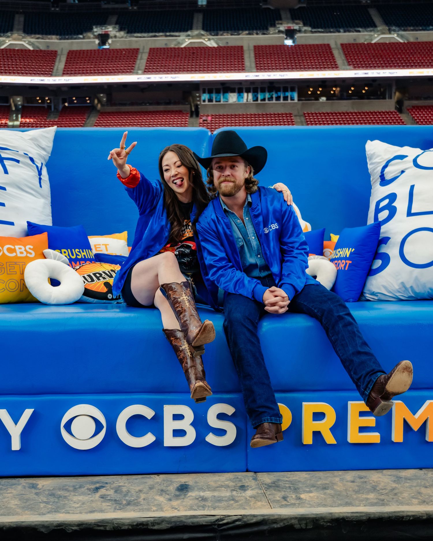 Two people sitting on a large blue CBS promotional couch in a stadium setting.