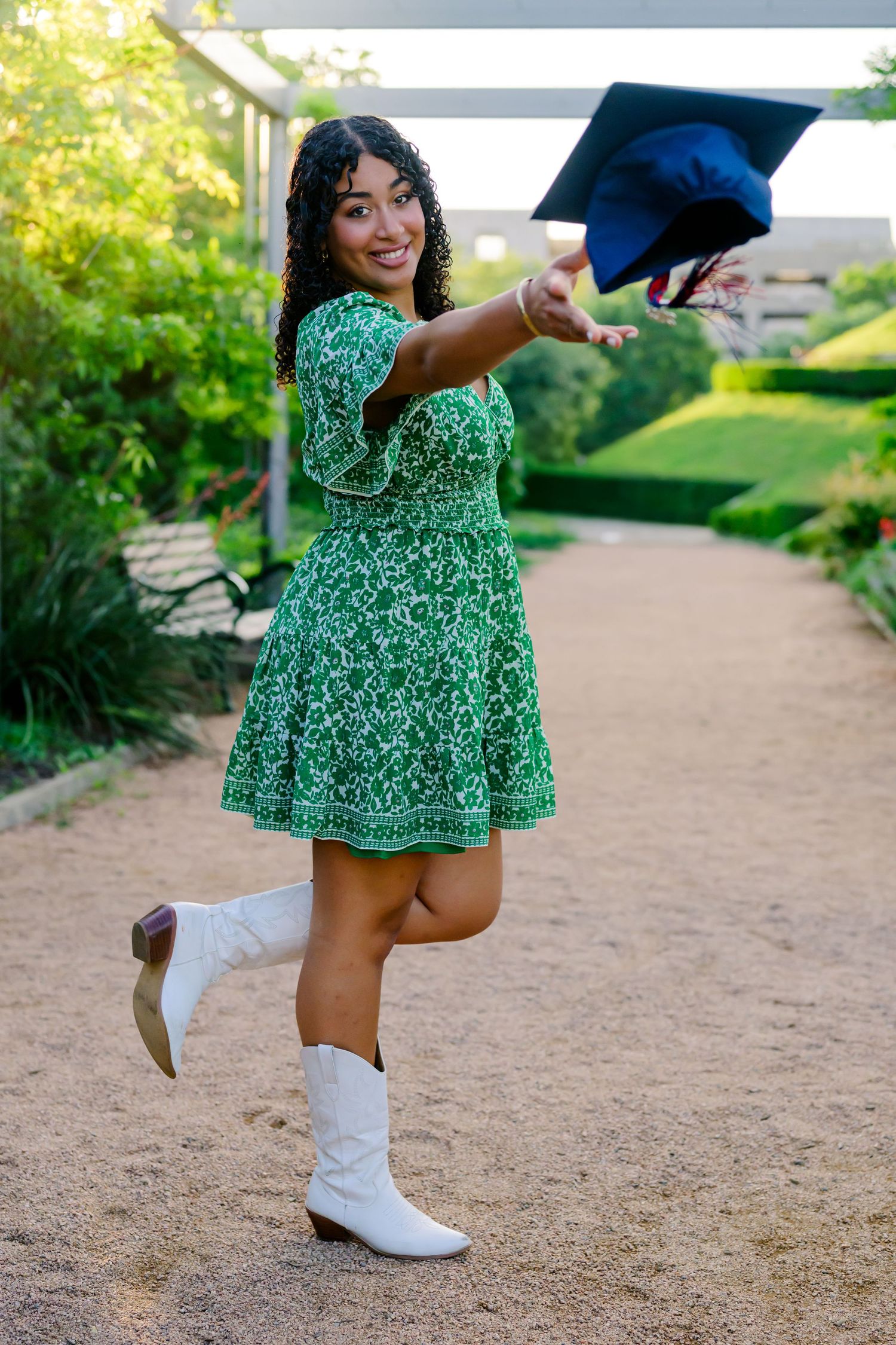 Graduate in green floral dress and white boots poses with graduation cap while celebrating outdoors.