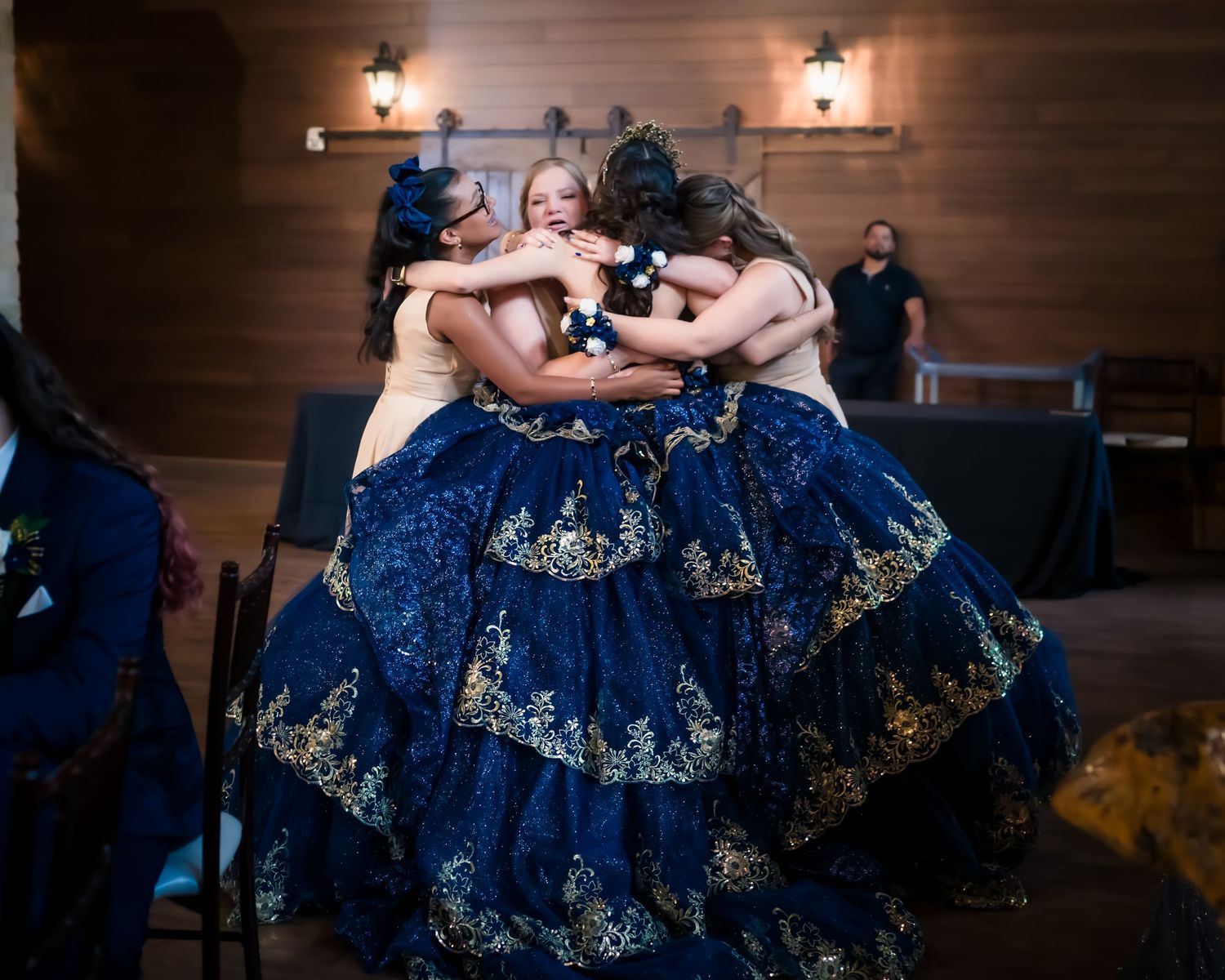 Dancers in navy blue quinceañera dresses with gold embroidery embrace in a joyful group hug during celebration.