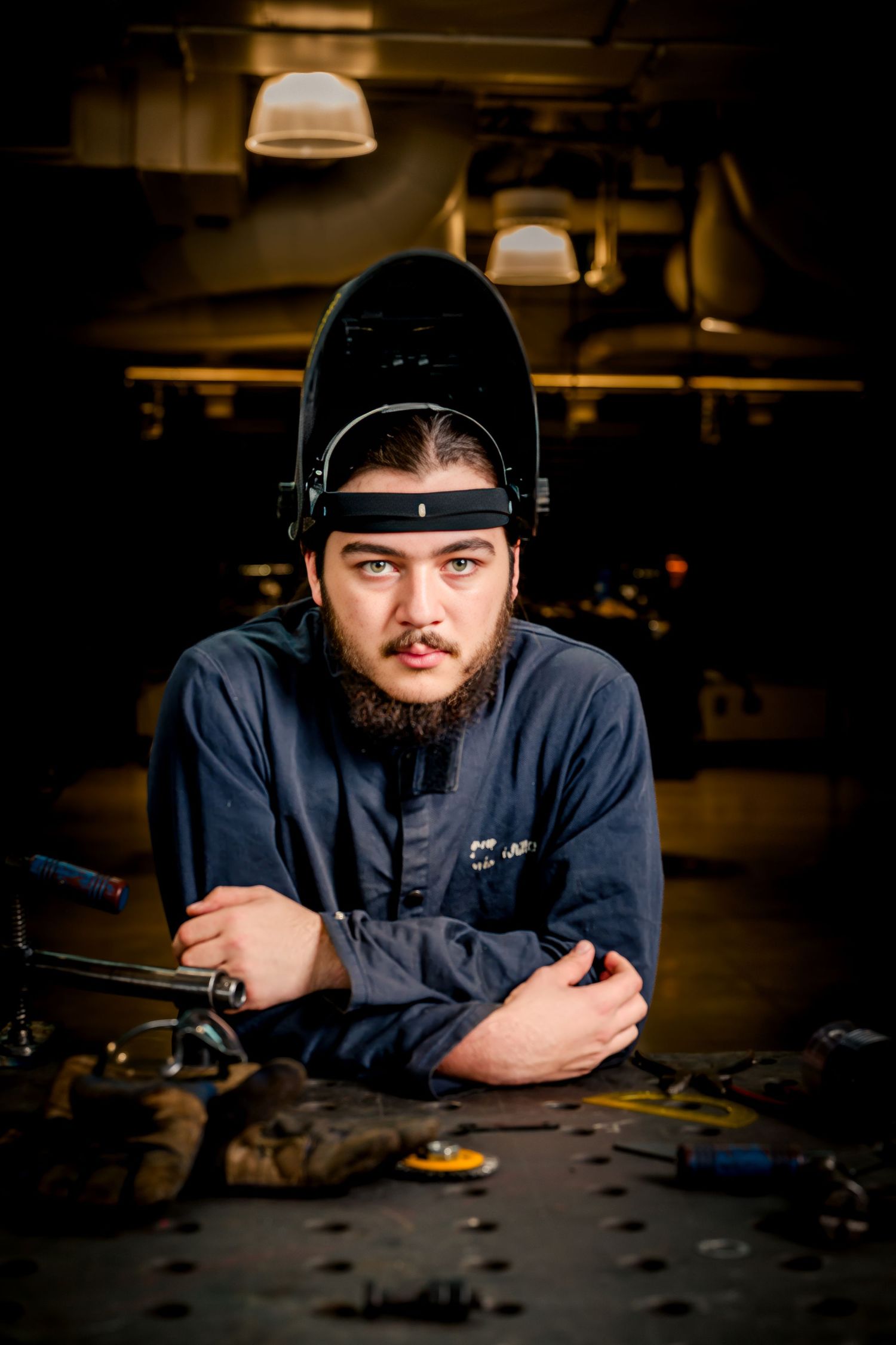 Young bearded welder with flipped-up welding mask, arms crossed at workbench in industrial workshop.