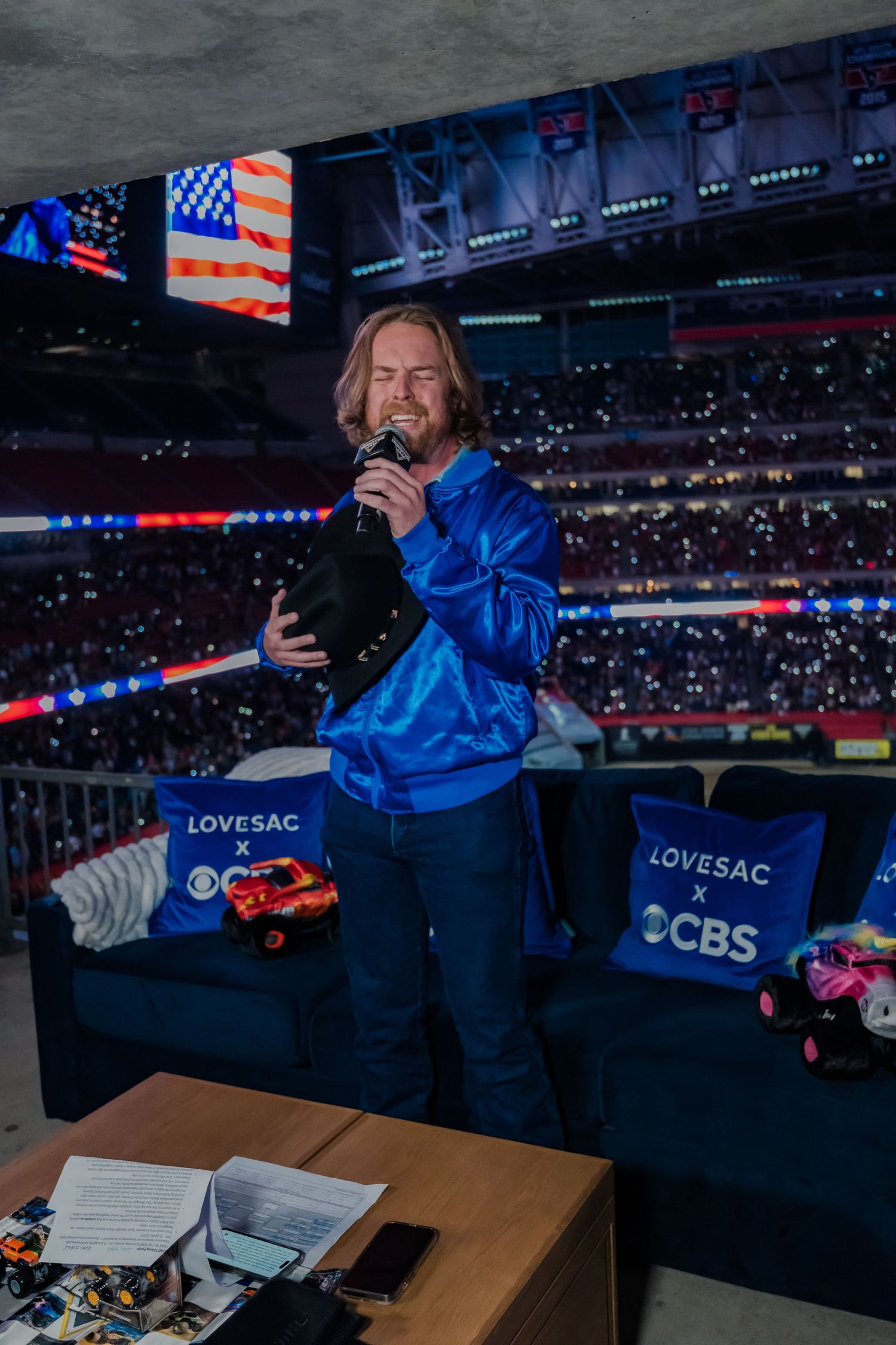 Sports commentator in blue jacket works at broadcast desk during game with American flag visible in stadium background.