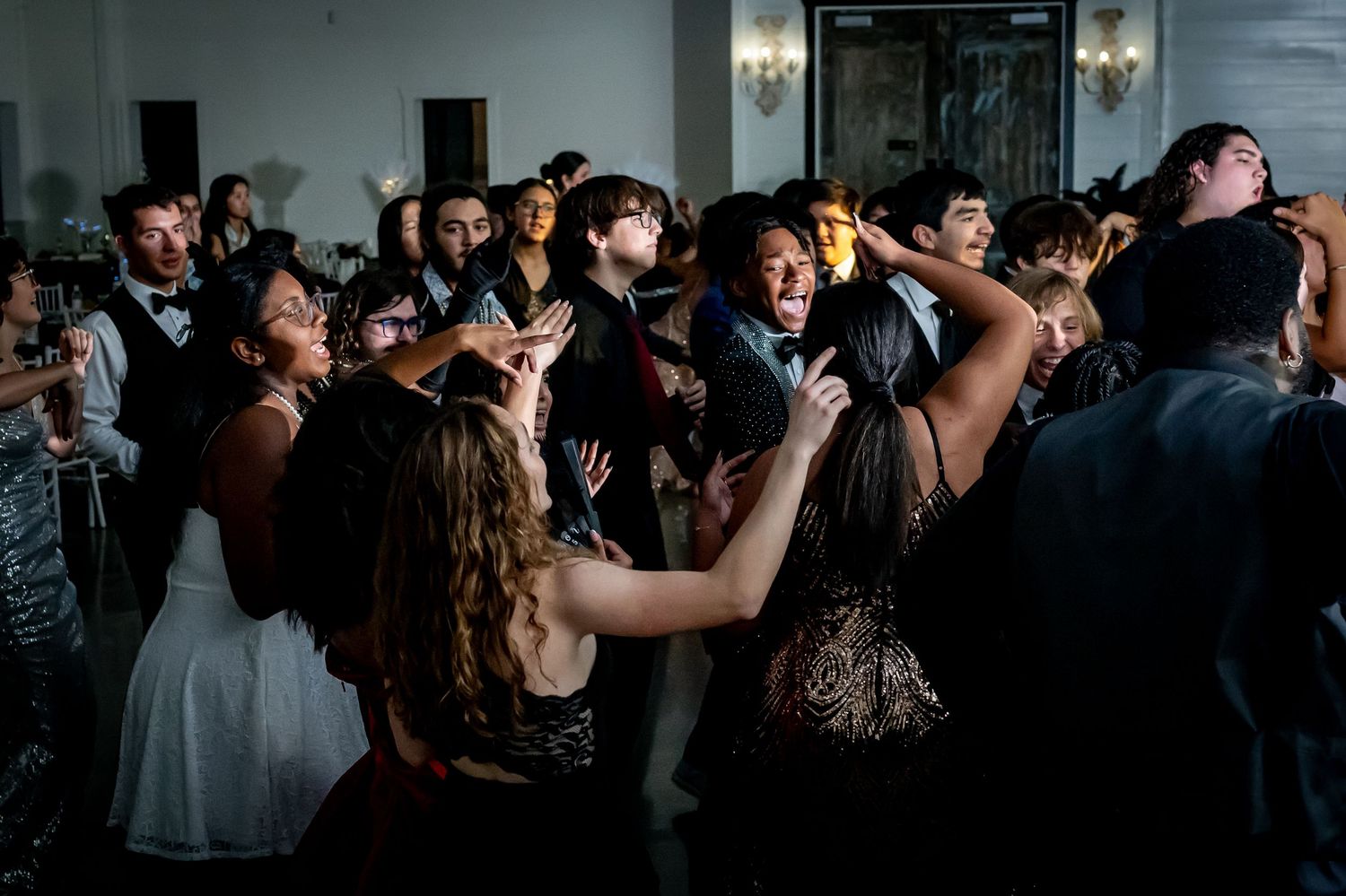 Energetic crowd dancing and celebrating at a wedding reception in a dimly lit ballroom with wall sconces.