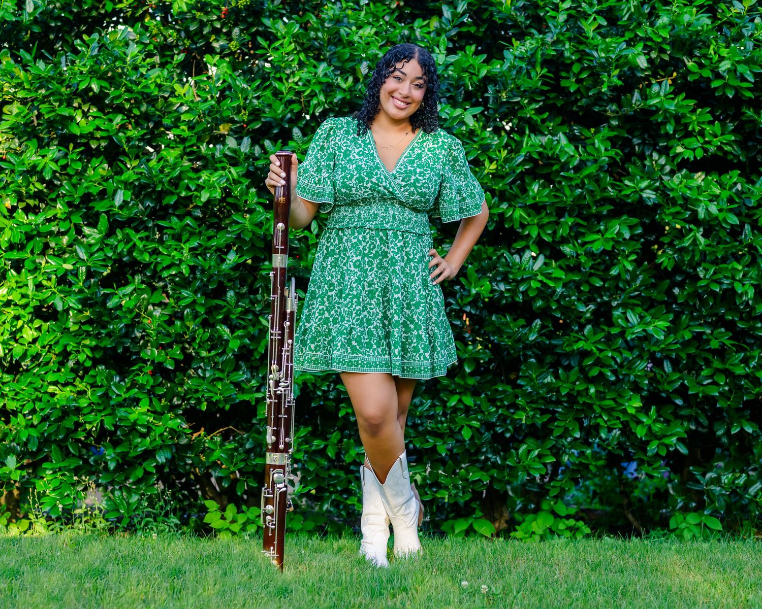 Someone in a green polka dot dress and white boots stands with their instrument against a lush green bush backdrop.