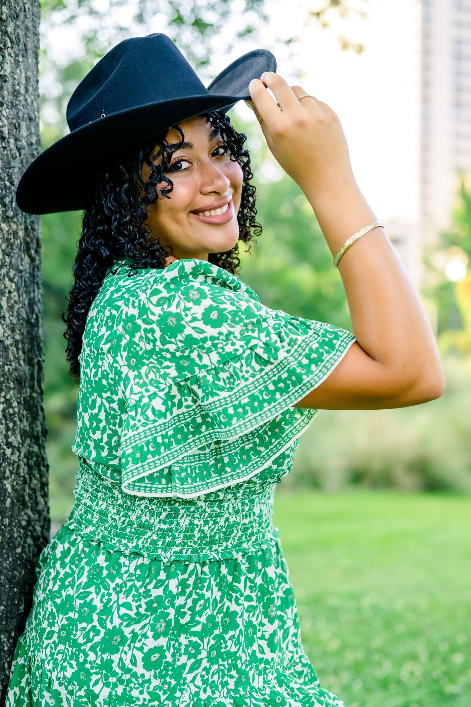 A person in a black cowboy hat and green floral dress poses against a tree in an outdoor setting.