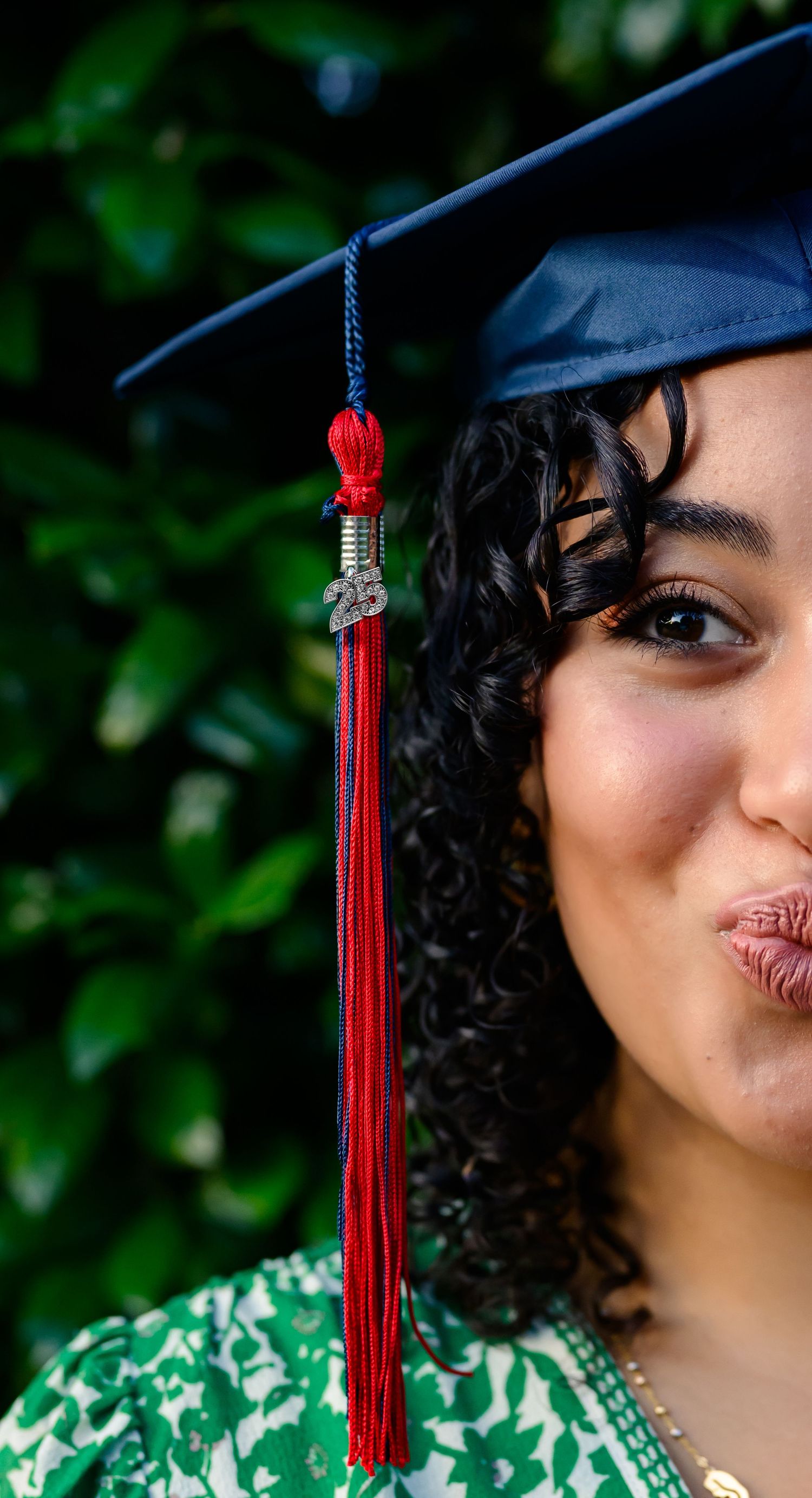Graduate in cap with red tassel poses with playful expression against green foliage background.
