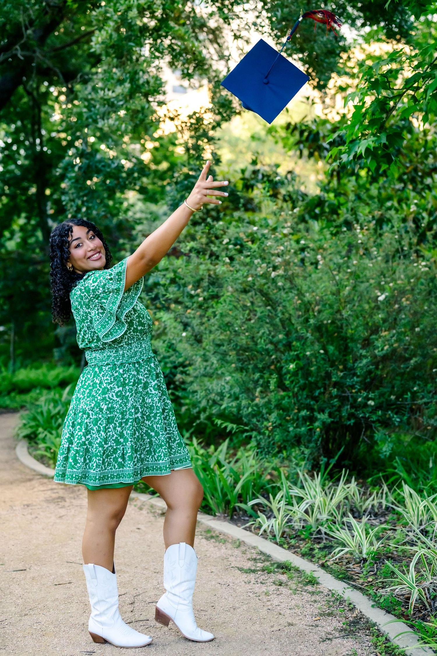 Graduate in green dress and white cowboy boots tossing blue cap in celebration outdoors near garden path.
