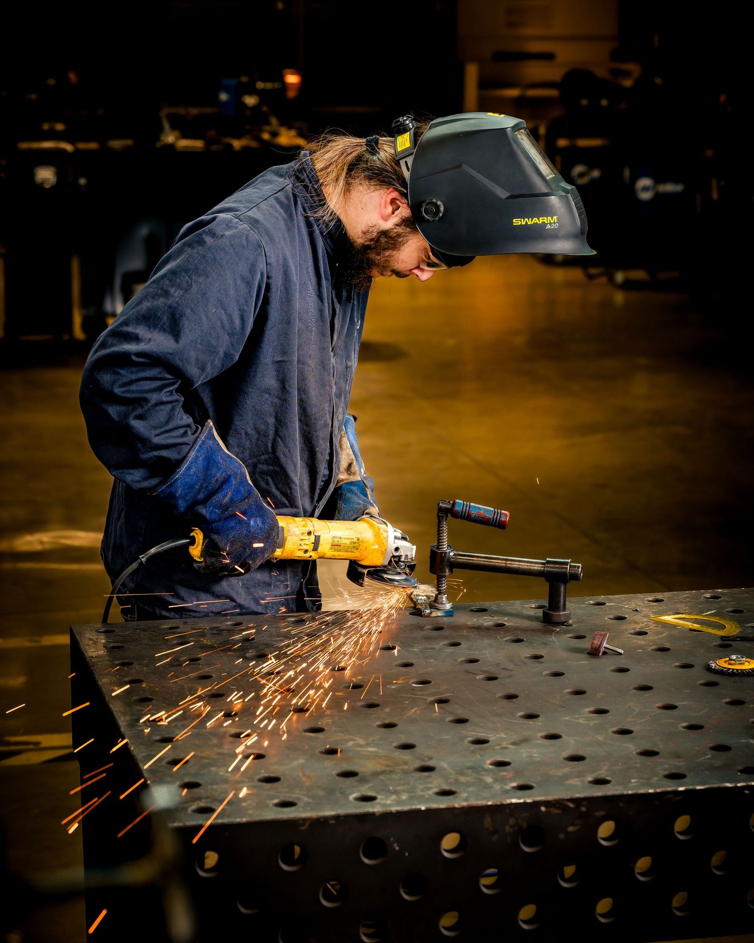 Welder using an angle grinder on a metal table, sending sparks flying, wearing a welding helmet and blue work jacket.