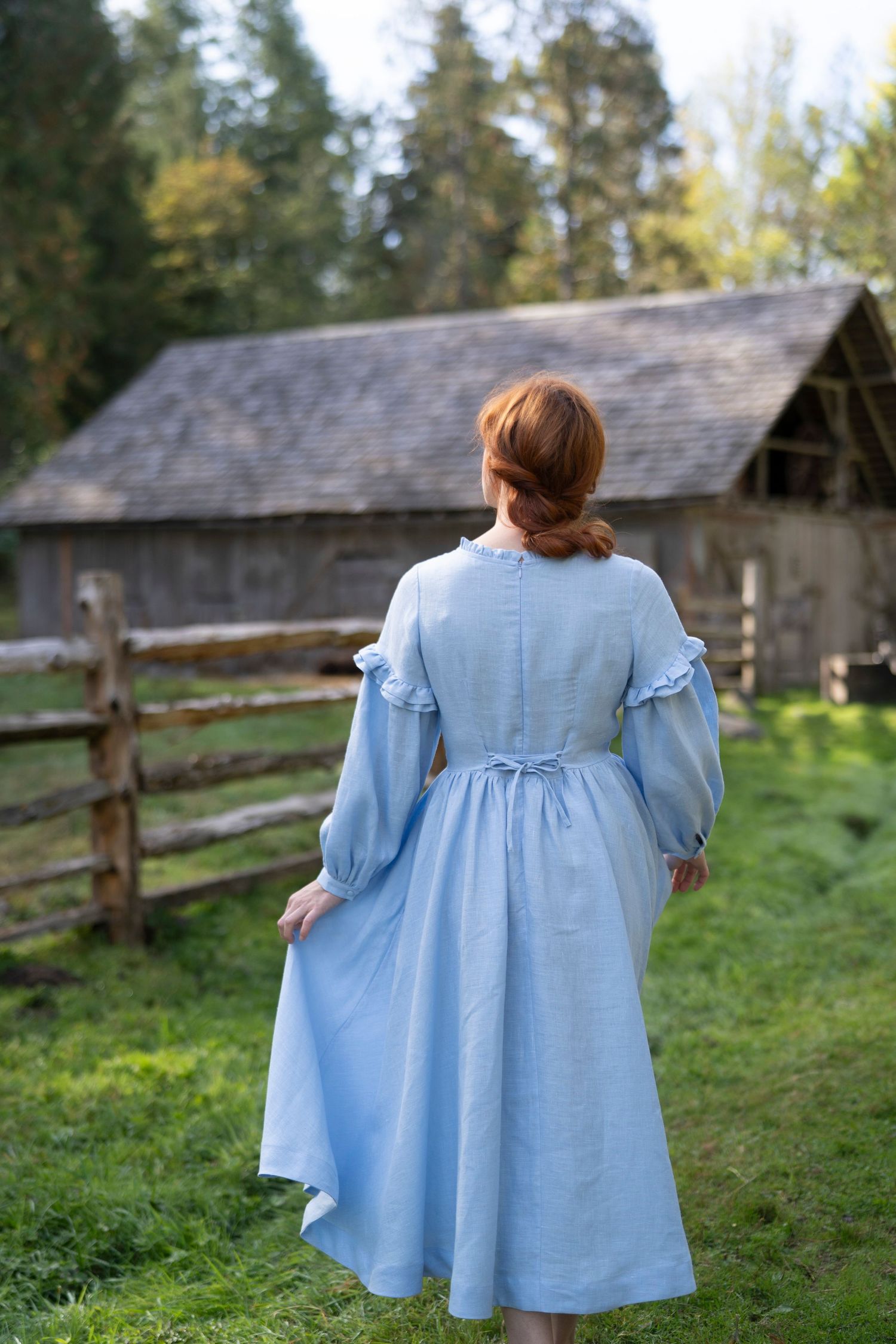 A person in a light blue vintage-style dress stands in front of a rustic barn and wooden fence.