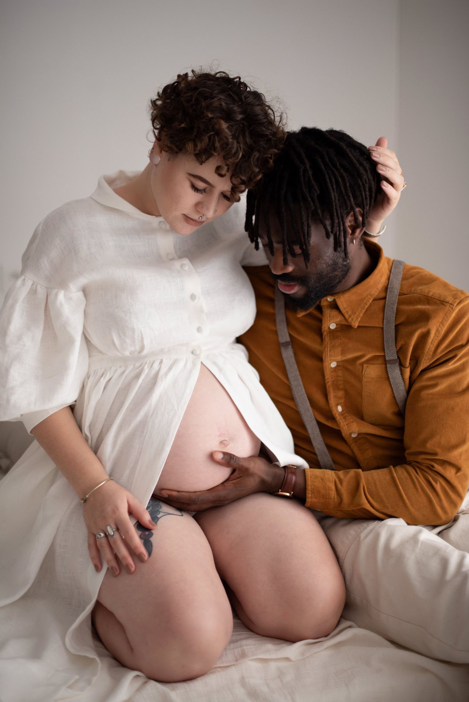 Maternity photo of couple in white dress and tan shirt touching belly against neutral background.