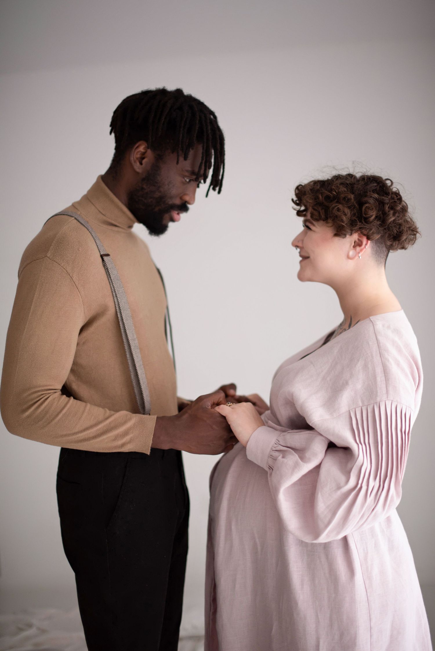 Couple holds hands during maternity shoot, wearing contrasting tan and pink outfits against neutral backdrop.
