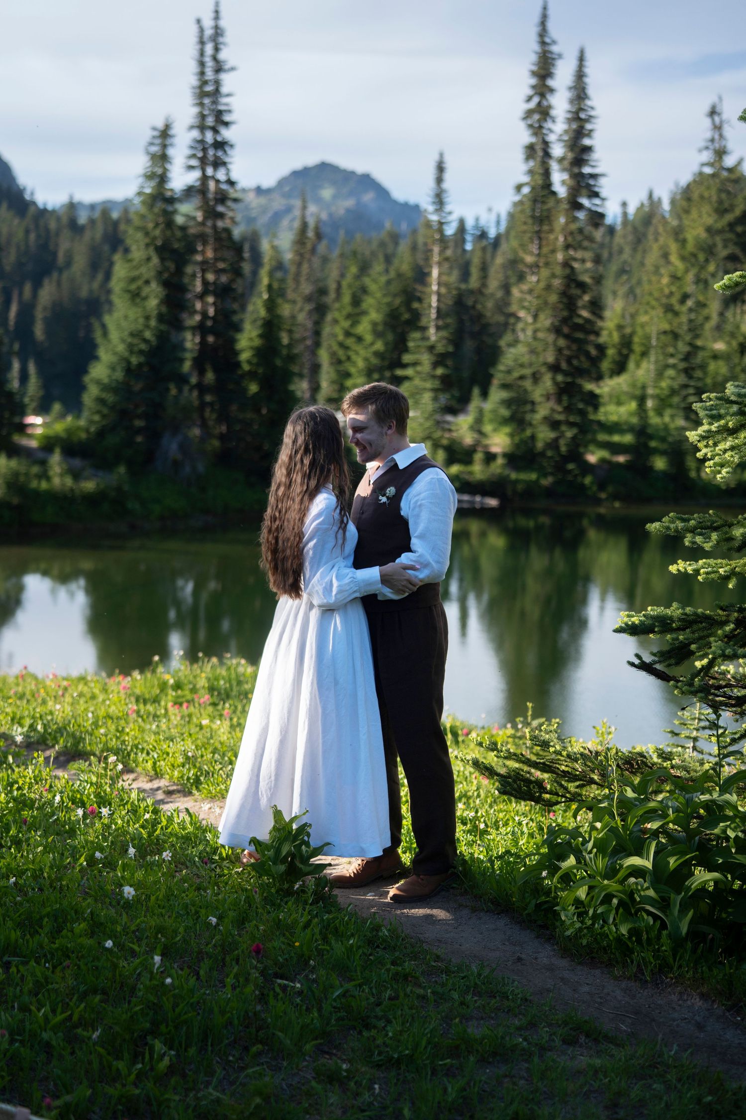 A couple shares a romantic moment by a serene mountain lake surrounded by evergreen trees and wildflowers at sunset.