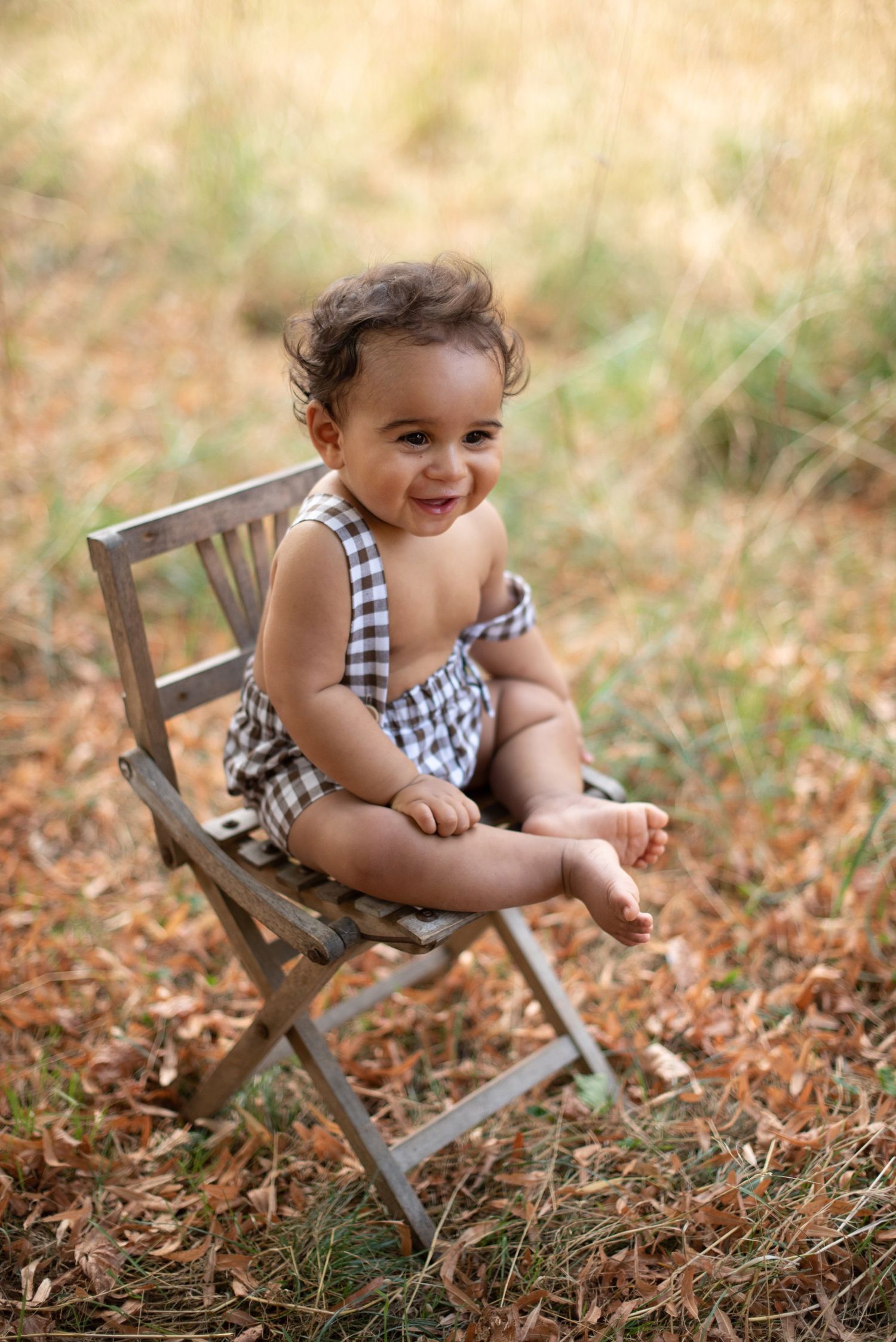A happy toddler wearing a gingham outfit sits on a wooden folding chair outdoors in autumn.