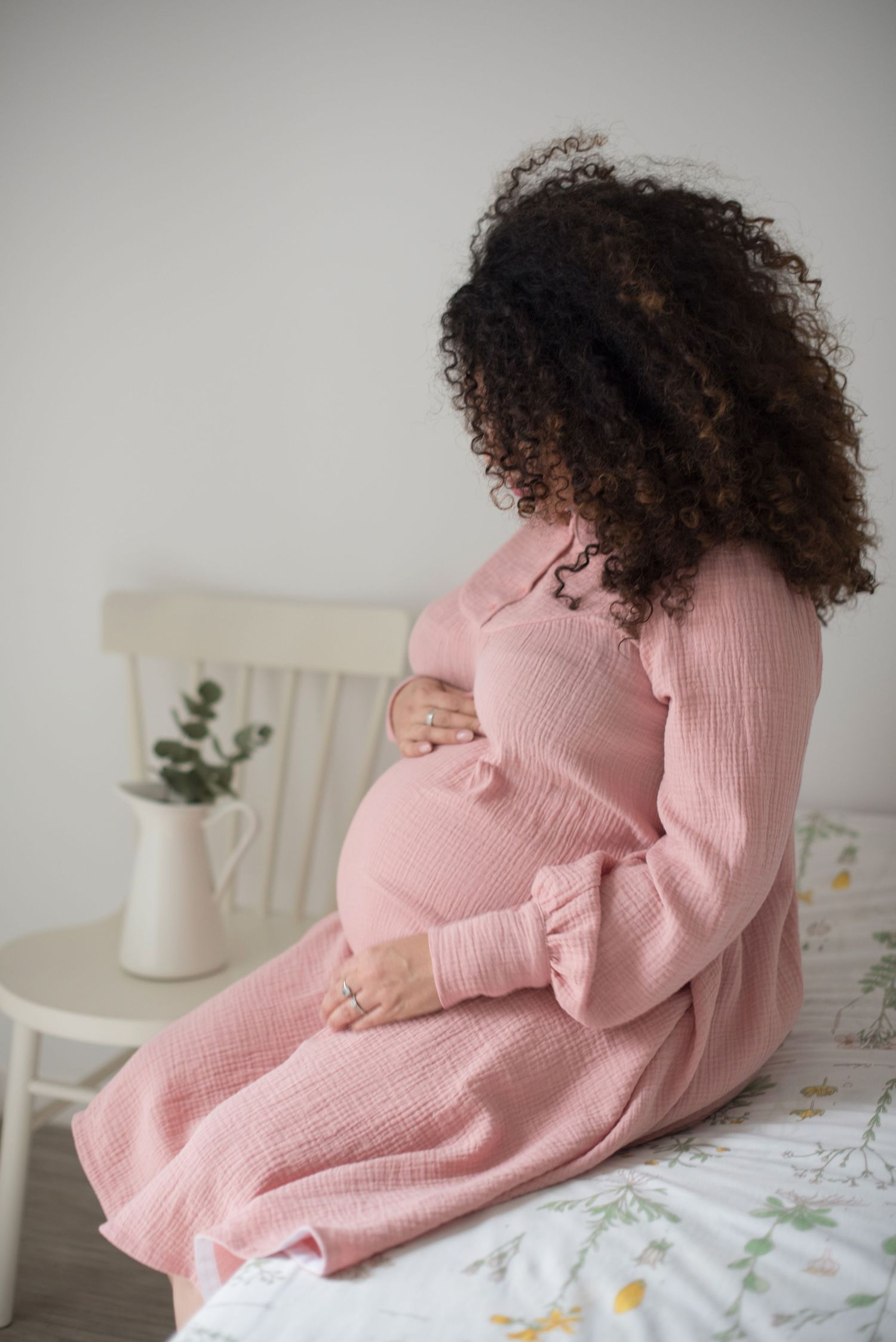 A pregnant woman in a pink dress sits on a bed, cradling her baby bump.