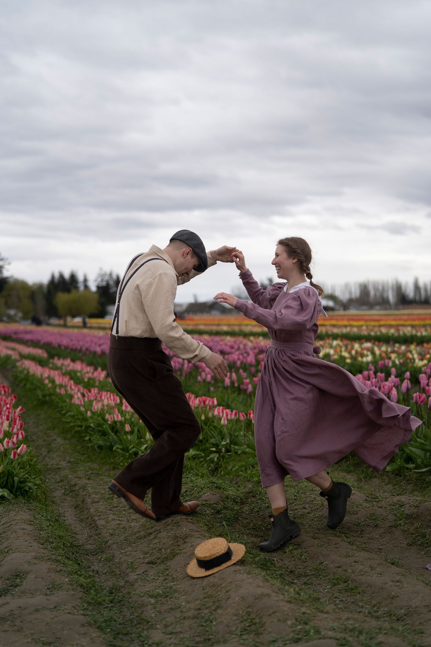 A couple dances together in vintage clothing among rows of colorful tulips on a cloudy day at a flower farm.