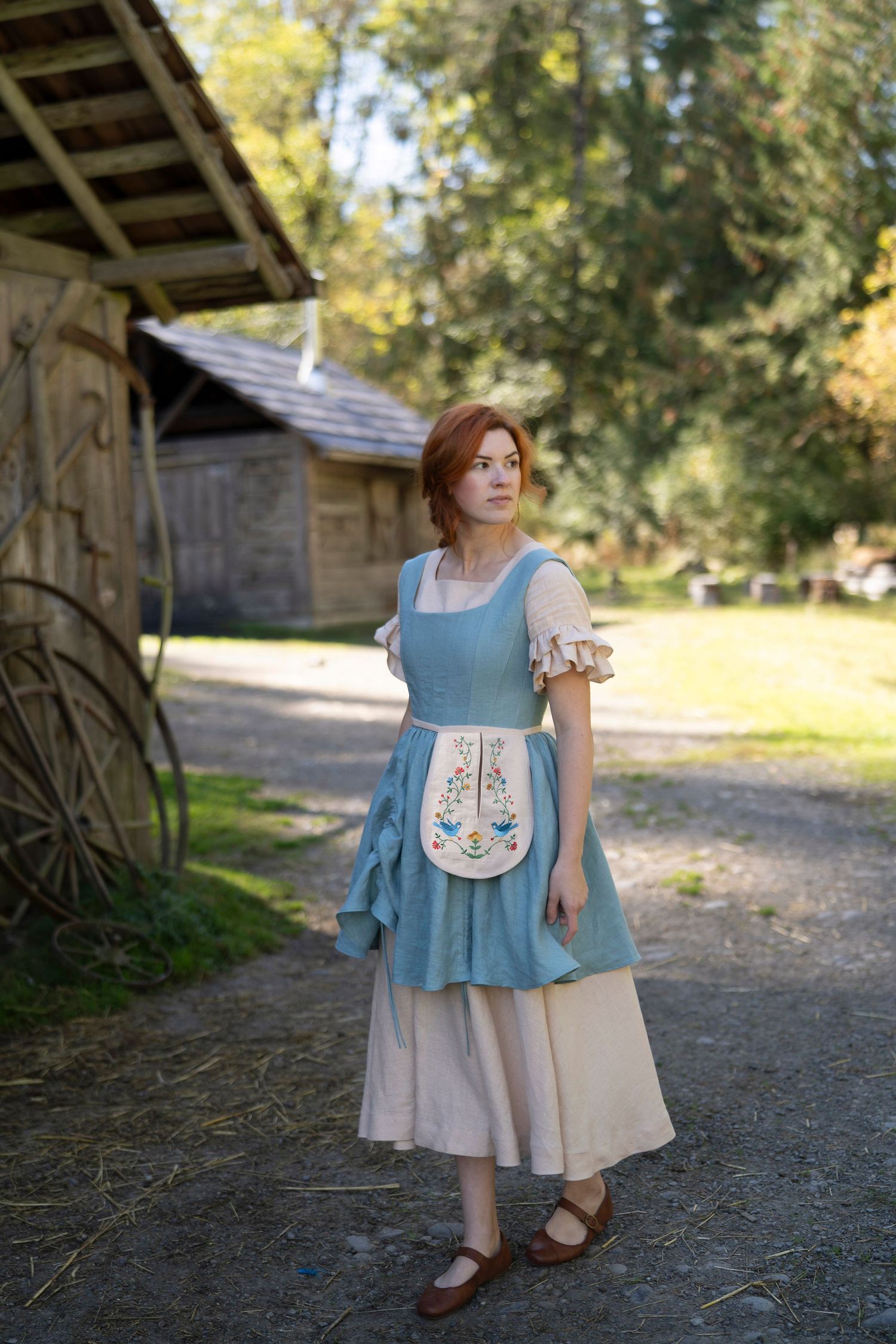 Woman wearing vintage blue pinafore dress and apron near rustic barn setting.