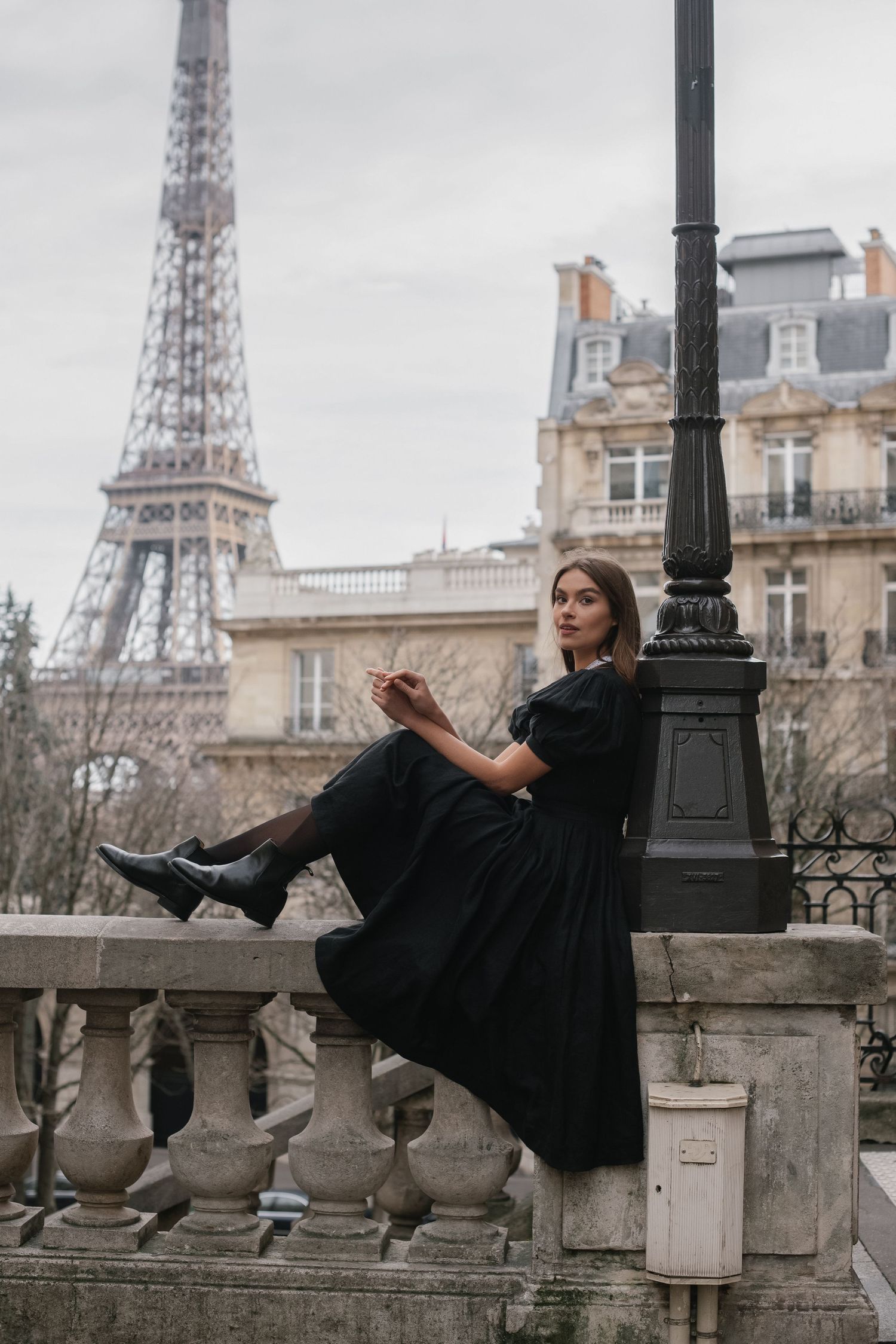 A woman in a black dress poses playfully on a stone balustrade with the Eiffel Tower in the background.