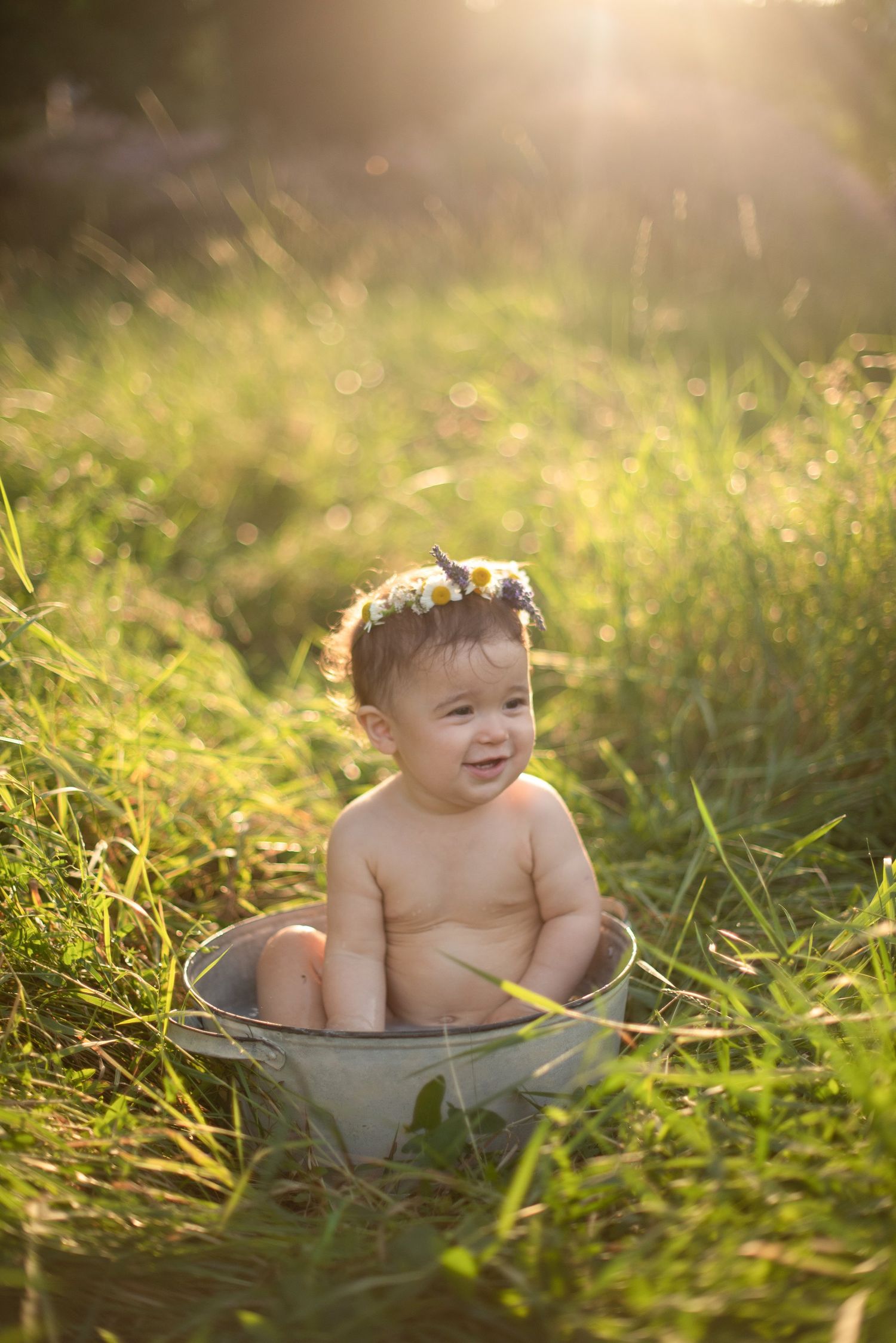 Happy baby sitting in metal tub surrounded by sunlit grass and flowers.