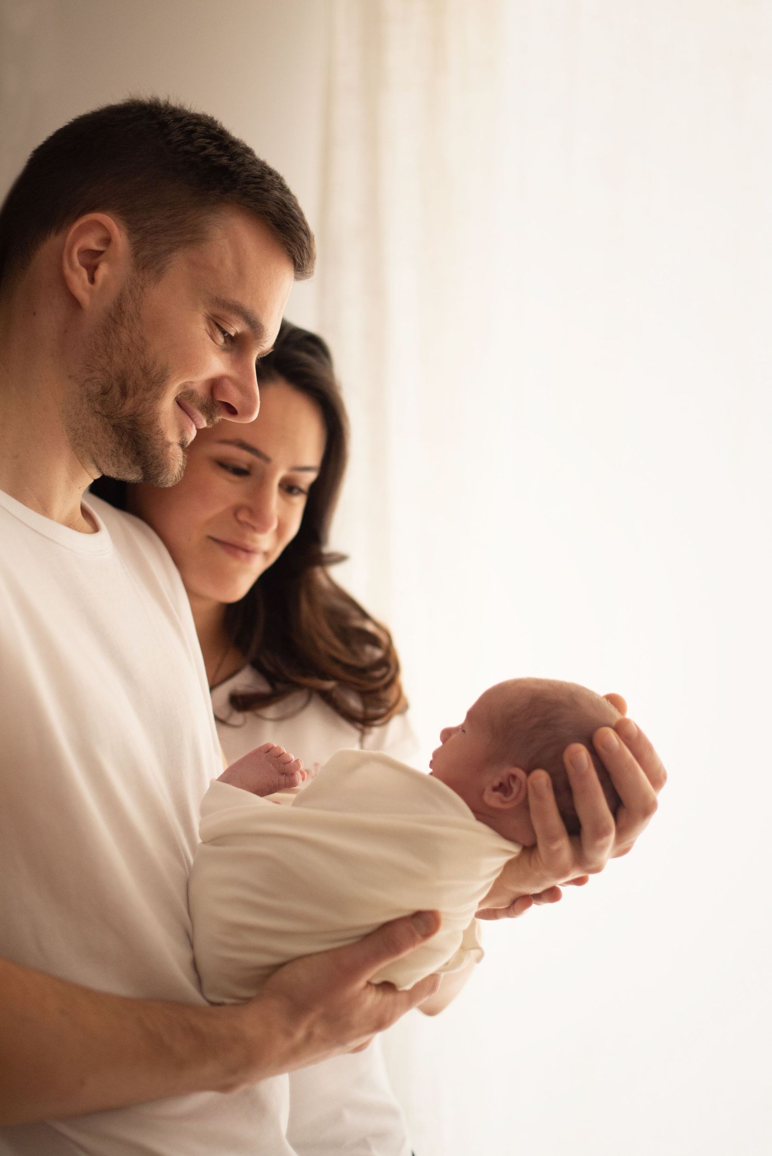 New parents share an intimate moment while holding their newborn baby in a tender embrace against soft lighting.