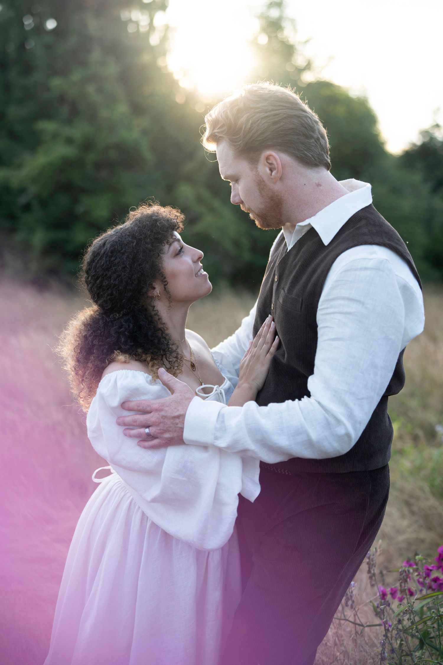 A romantic couple embraces in golden sunlight surrounded by pink wildflowers during a sunset photo session.