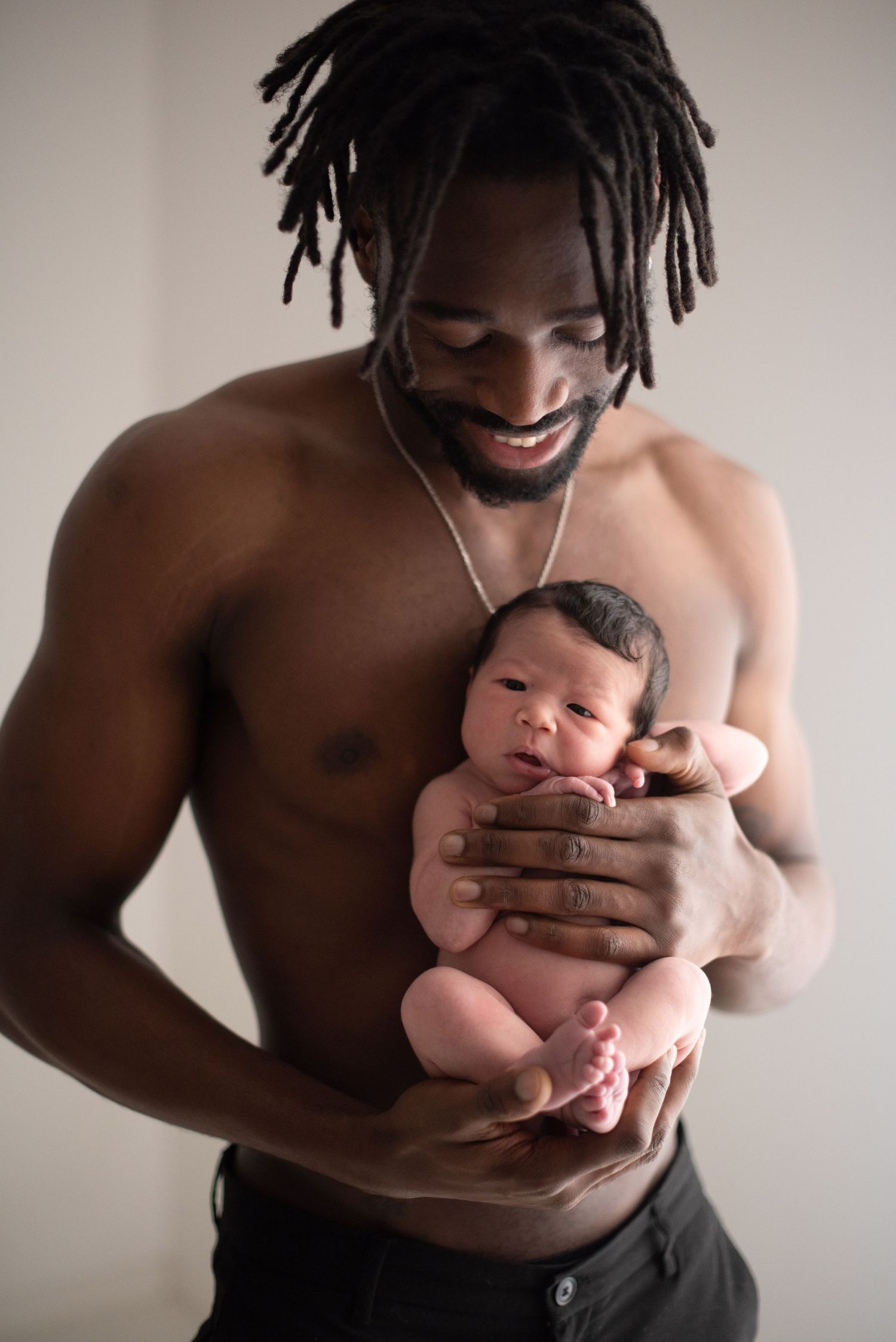 A shirtless man with dreadlocks tenderly holds a newborn baby against his chest in a studio portrait.