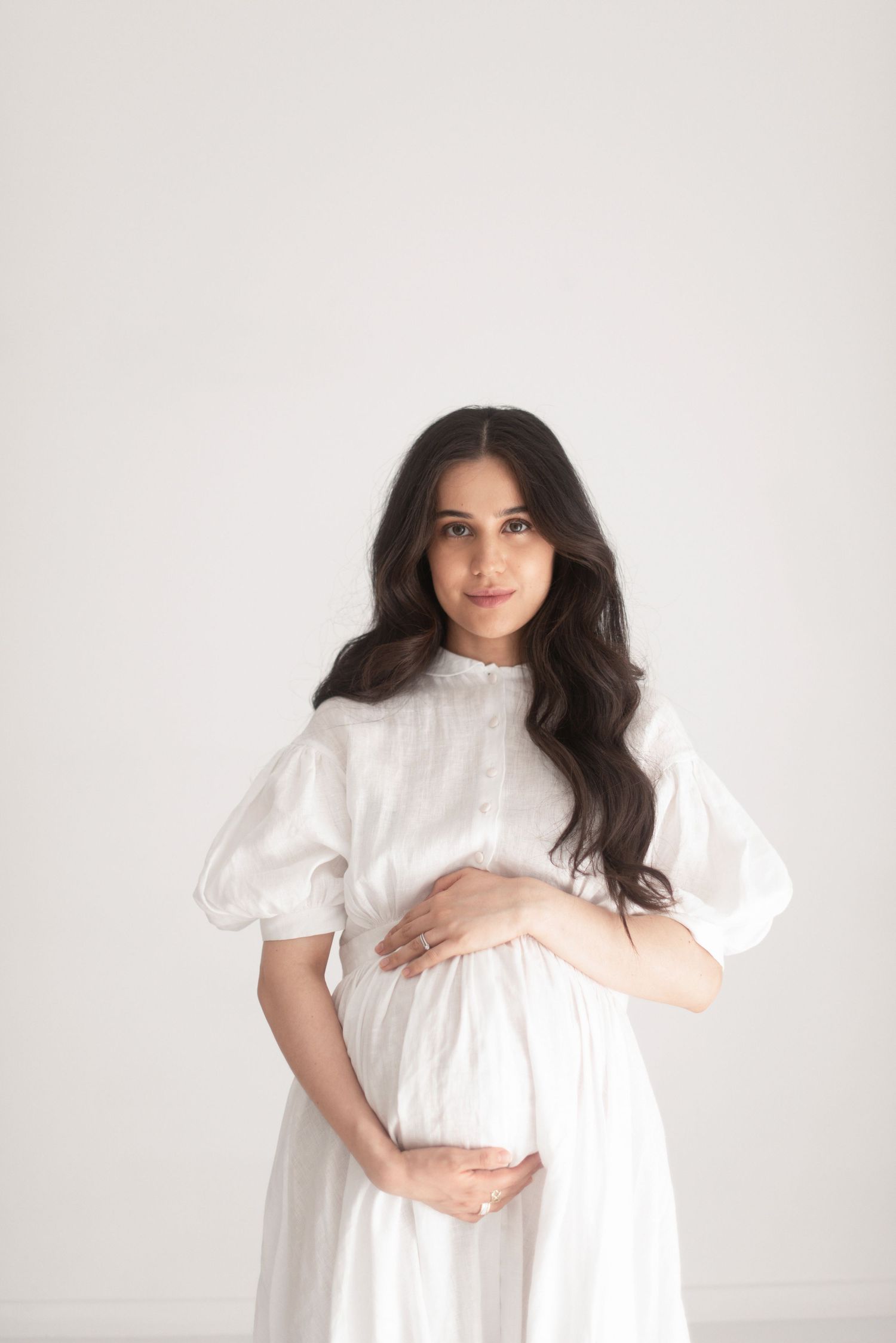 Elegant maternity portrait in white puffy-sleeved dress against neutral background.