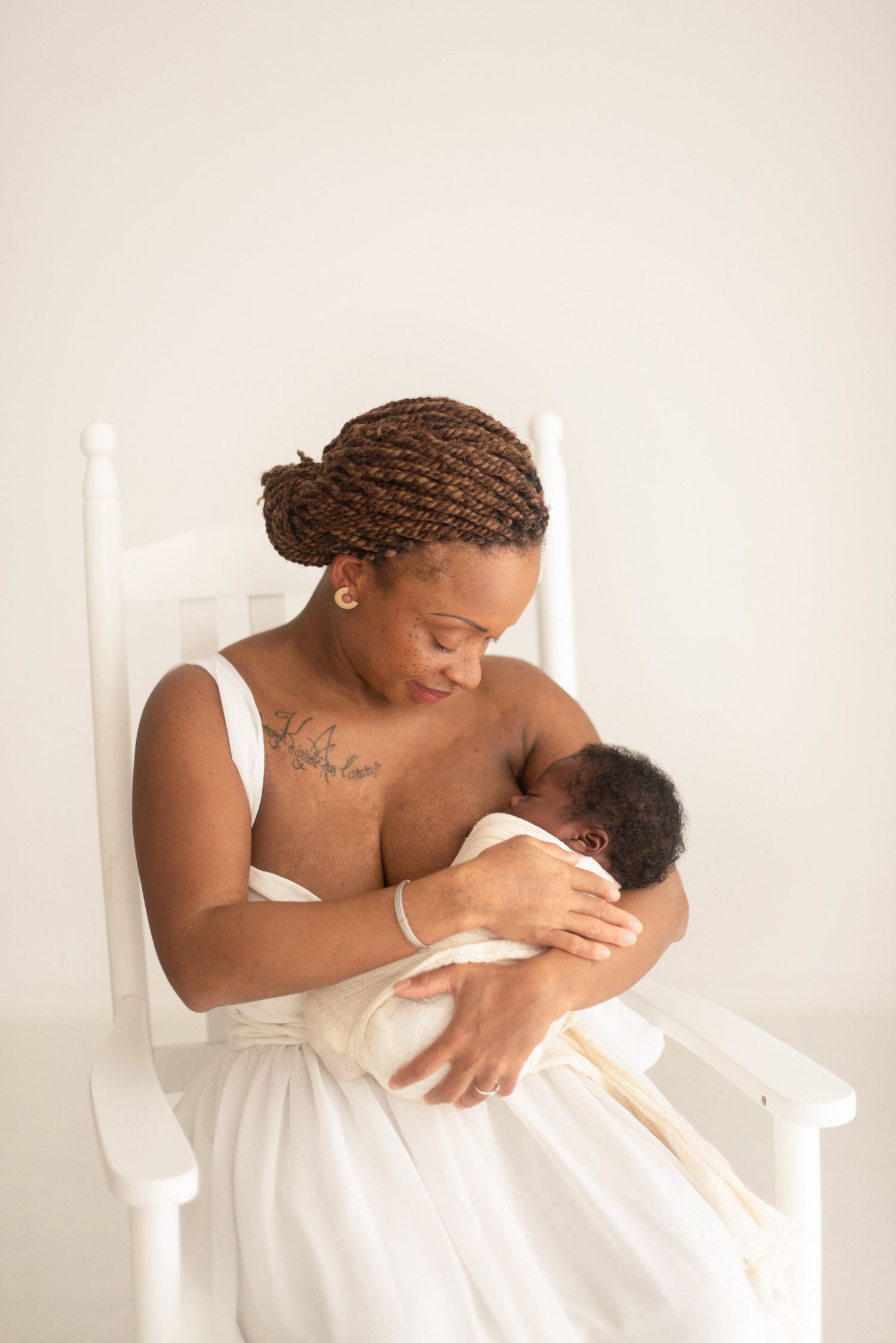 Mother in white dress tenderly holding and feeding newborn baby in white rocking chair.