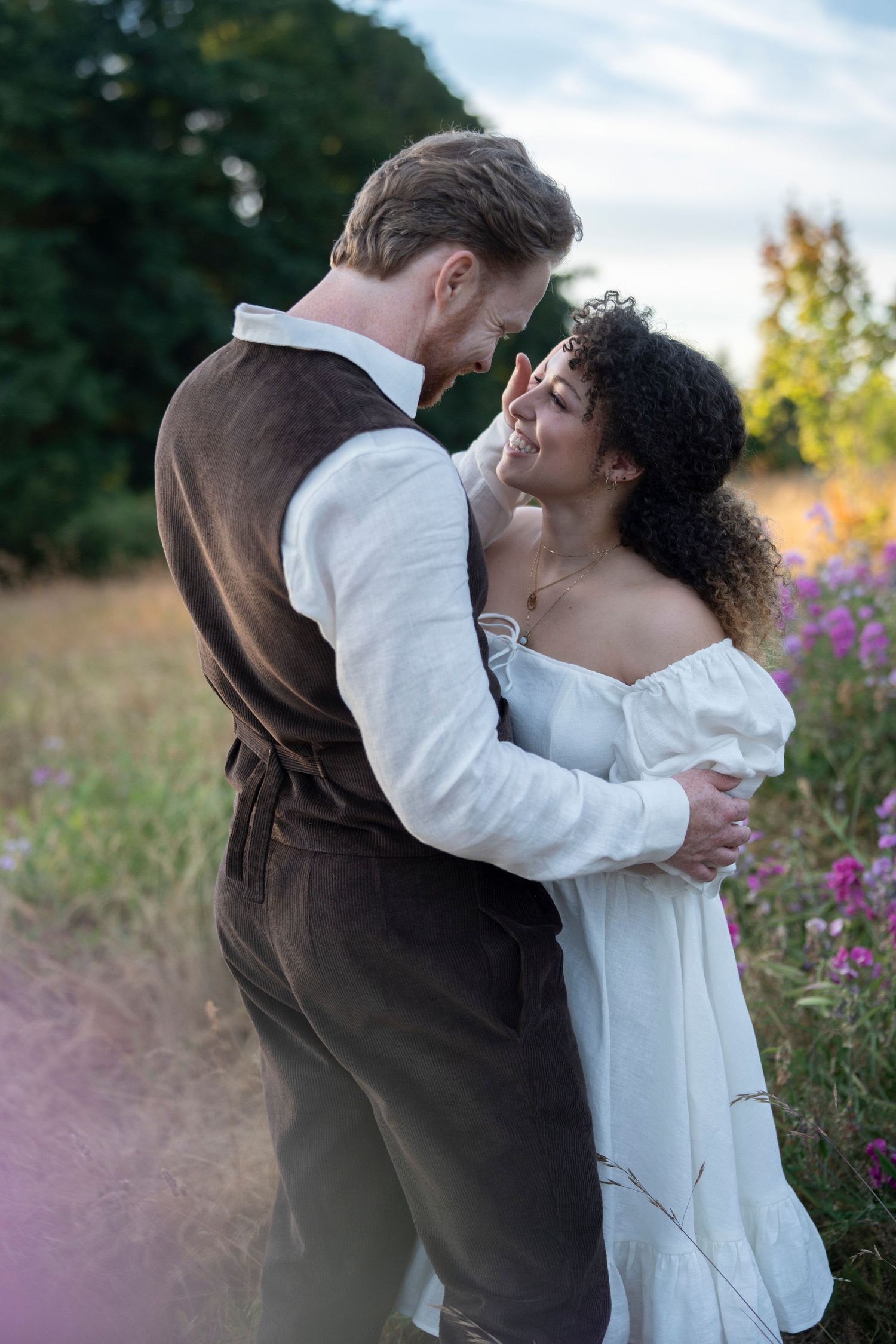 A couple in contrasting brown and white outfits share an intimate embrace in a wildflower meadow at sunset.
