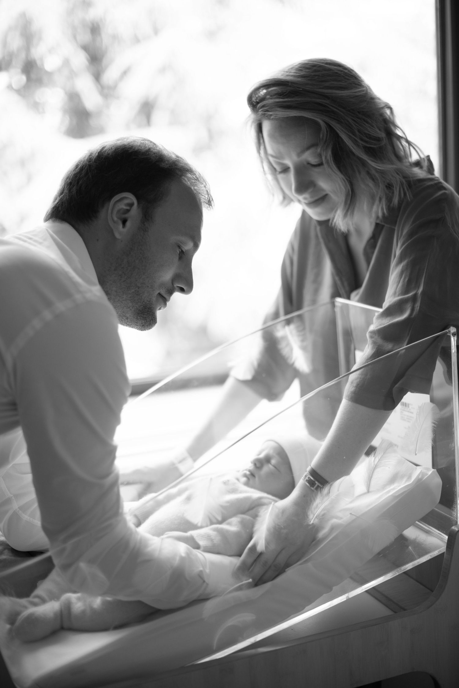A tender black and white moment between parents and their newborn baby in soft natural lighting.