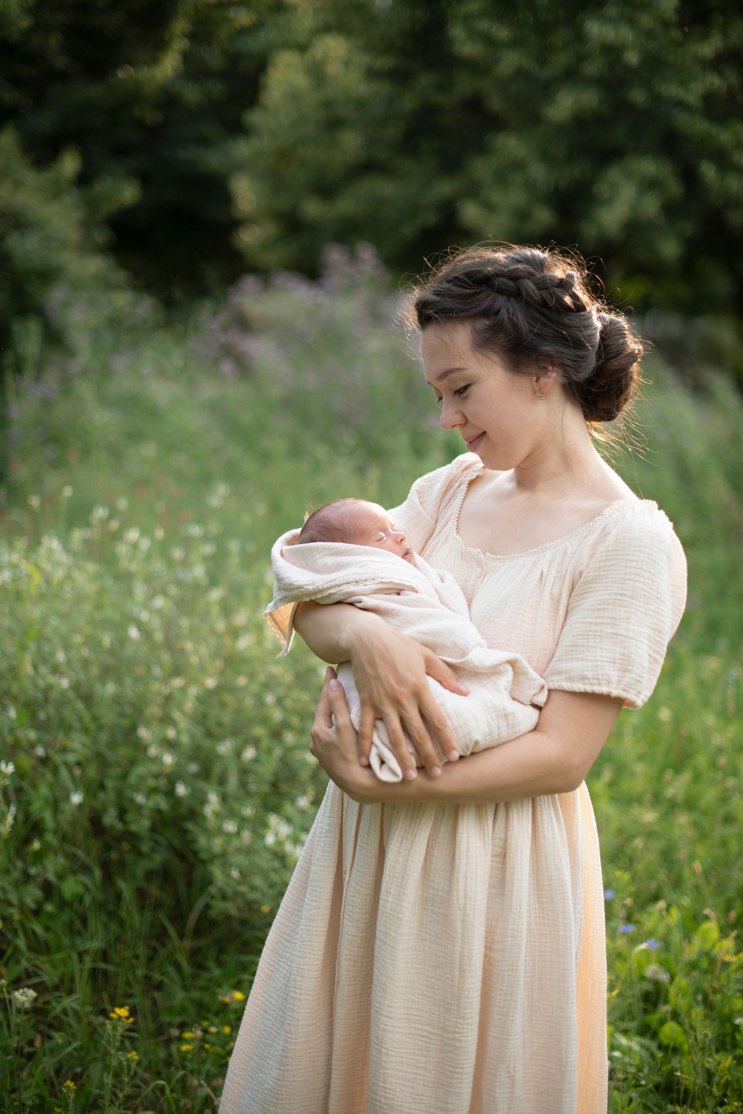 A woman in a cream-colored dress holds a newborn baby wrapped in white, standing in a sunlit meadow with wildflowers.